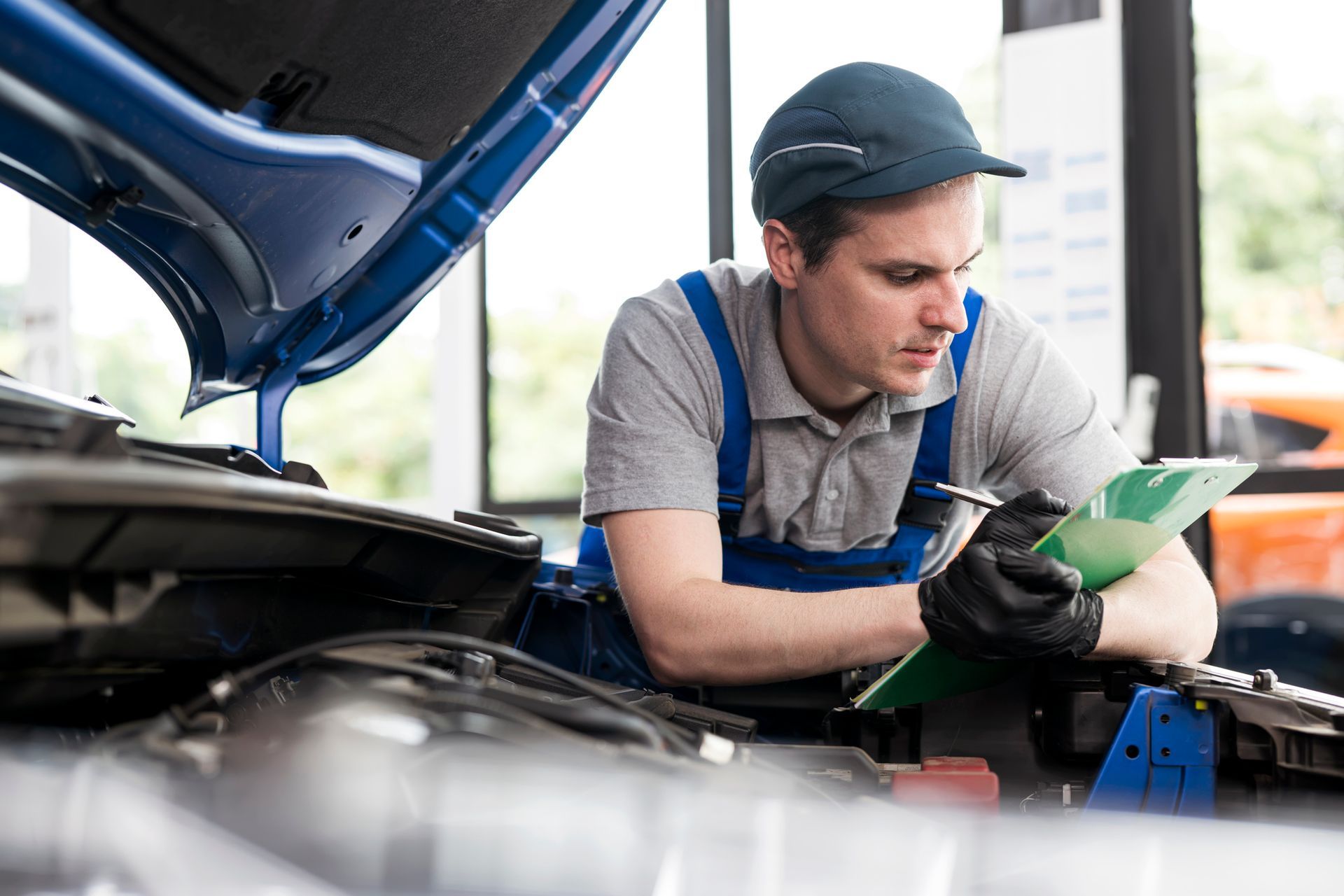 Car mechanic takes notes while checking a car’s hood, inside a car repair shop. Car mechanic takes notes while checking a car’s hood, inside a car repair shop.