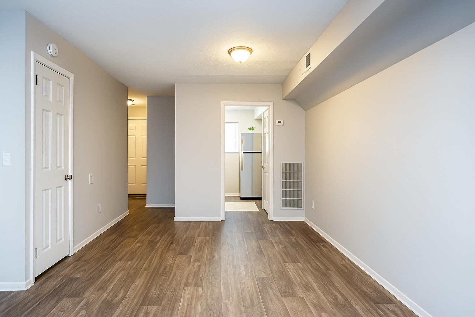Empty apartment interior with wooden floor, white walls, and doorway to the kitchen.