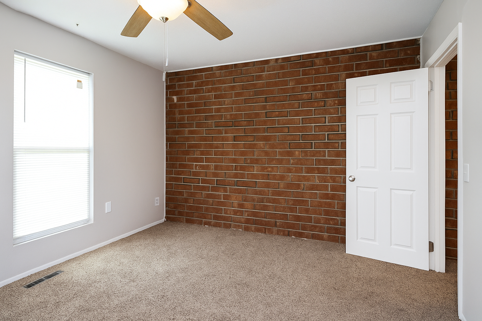Empty bedroom with brick accent wall, beige carpet, window with blinds, and open white door.
