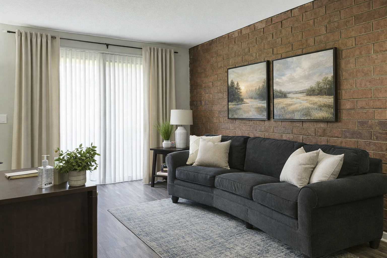 Living room with gray sofa, brick wall, and beige curtains. Two landscape paintings hang above the sofa.