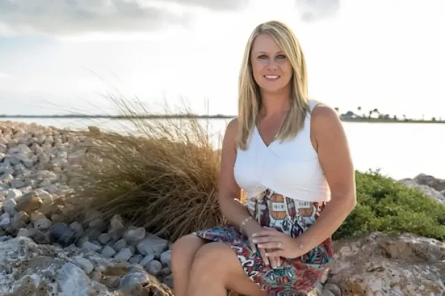 Woman sitting on rocks by a lakeshore, smiling in a white top and patterned skirt.