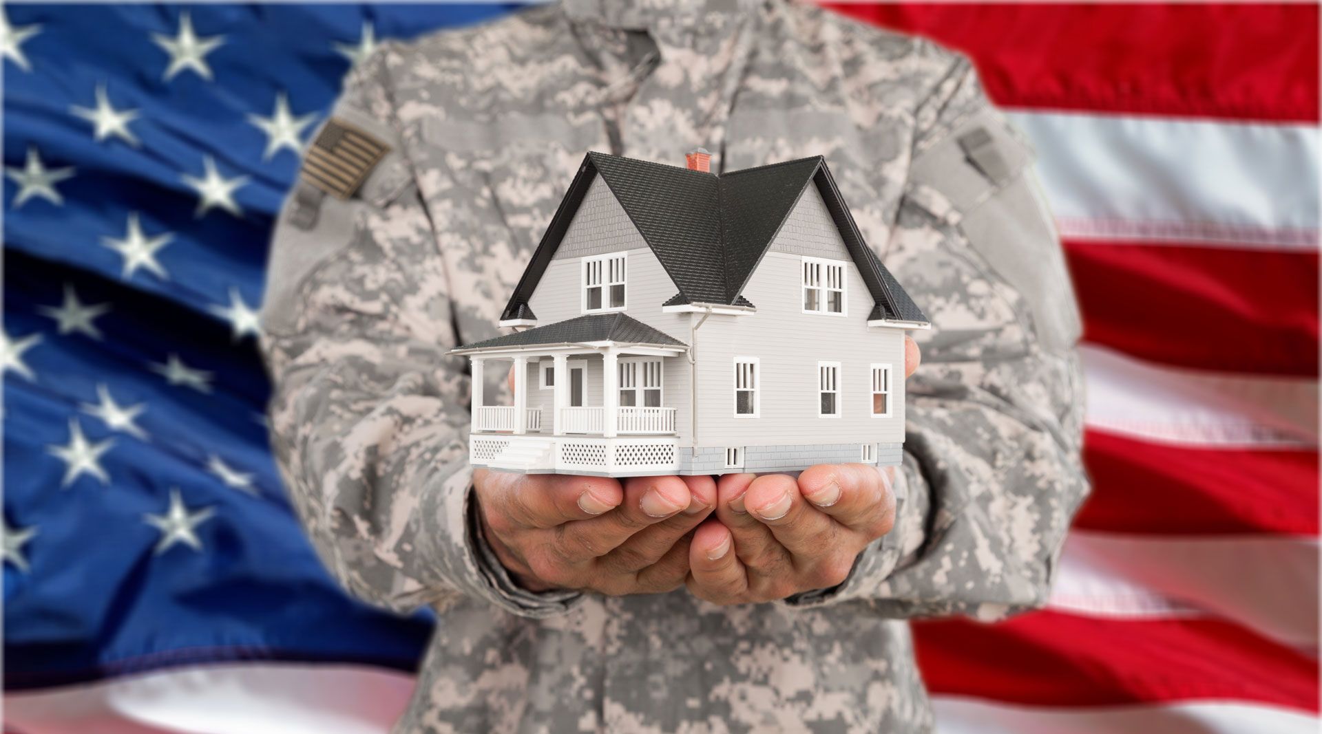 Soldier in camouflage holding a small white house with a U.S. flag in the background