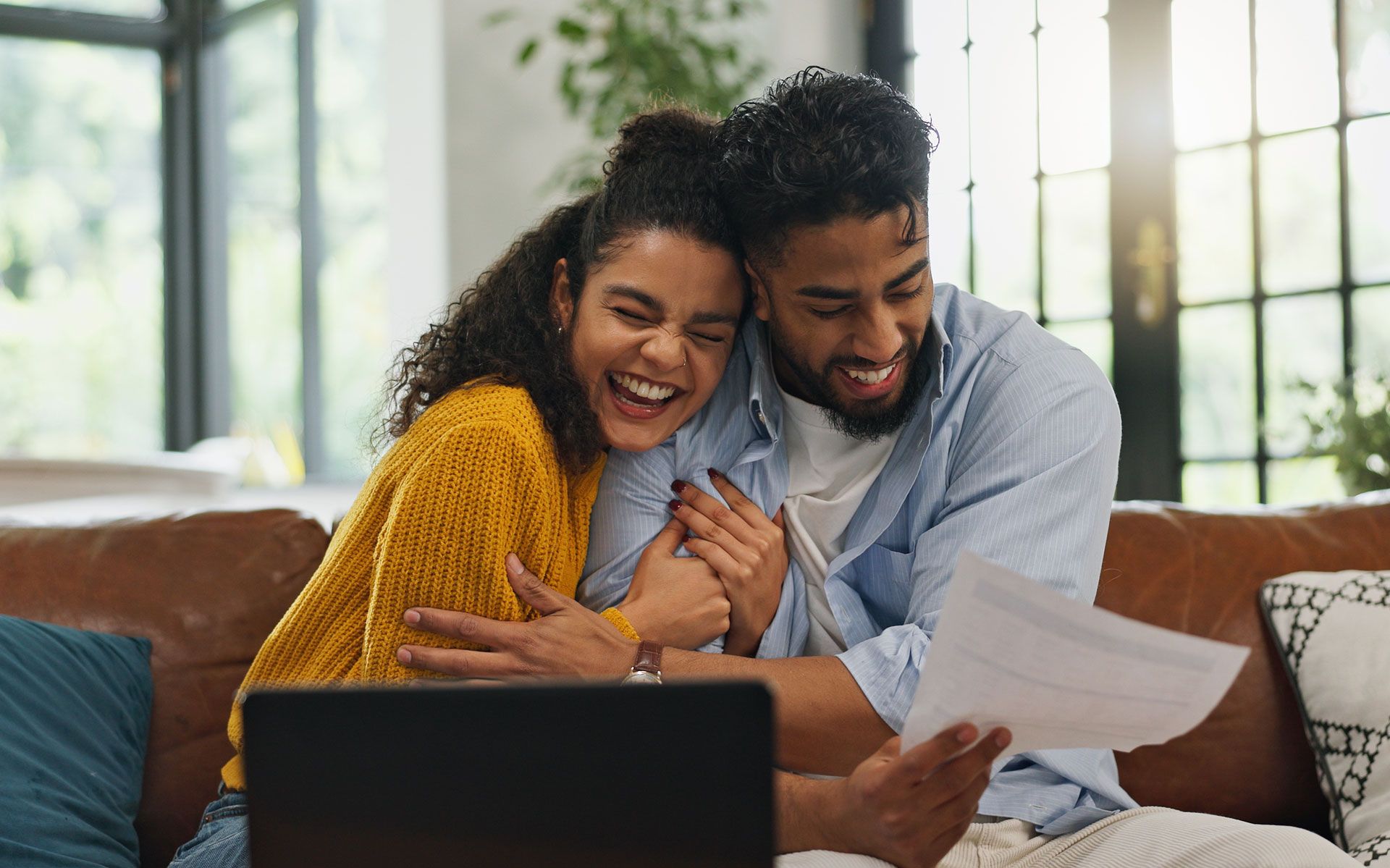 Couple smiling on a couch, reading papers together beside a laptop in a bright living room