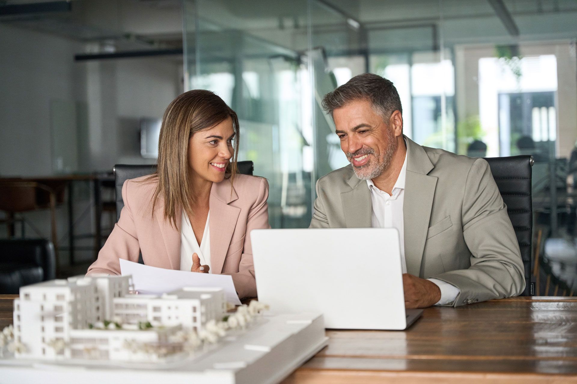 Two professionals review a laptop at a conference table with building models in the foreground.