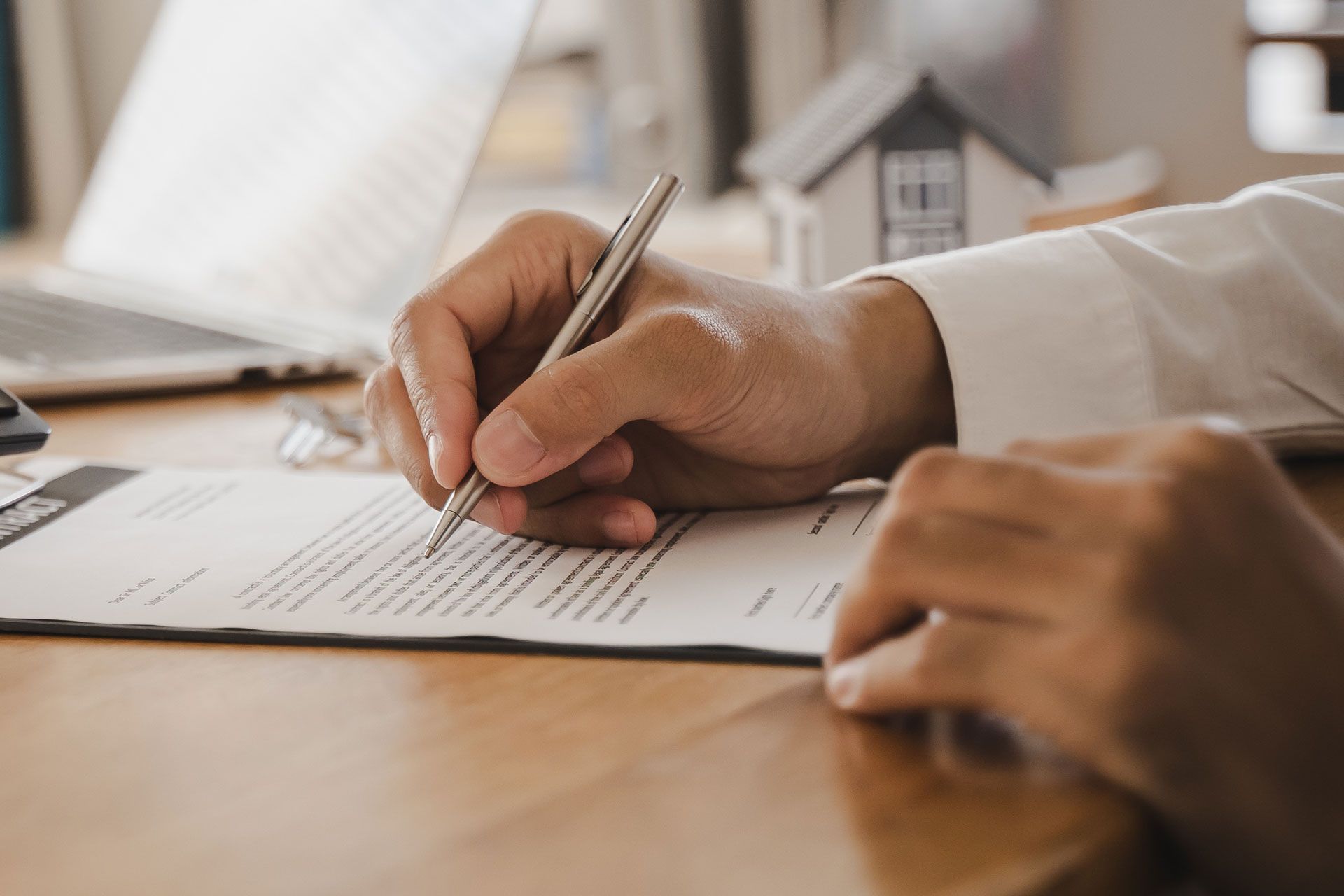 Hand signing a document at a wooden desk, with a laptop and house model in the background