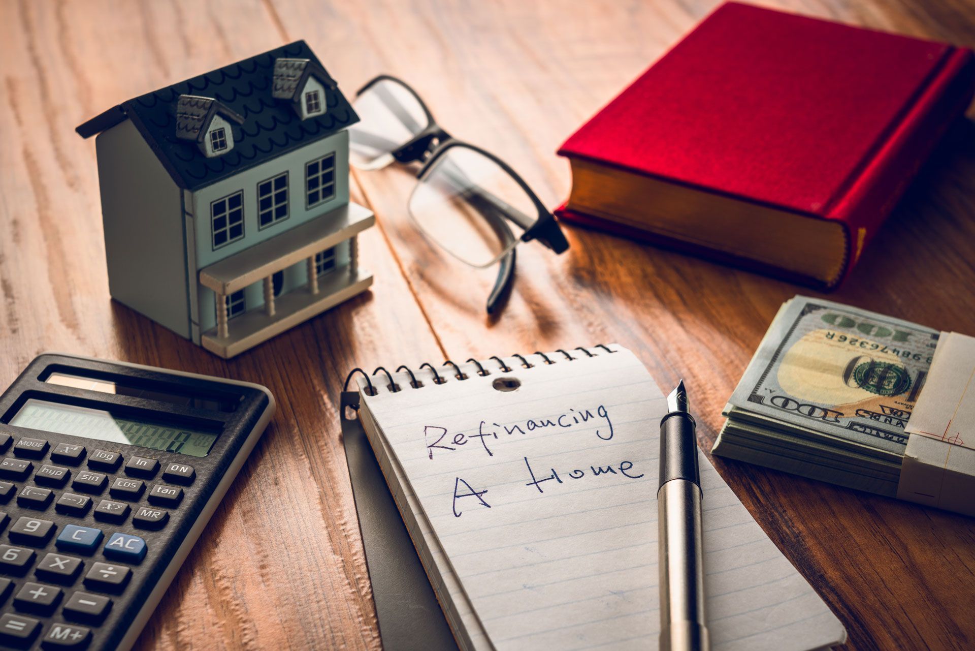 Calculator, toy house, eyeglasses, notebook, red book, and cash on a wooden desk.
