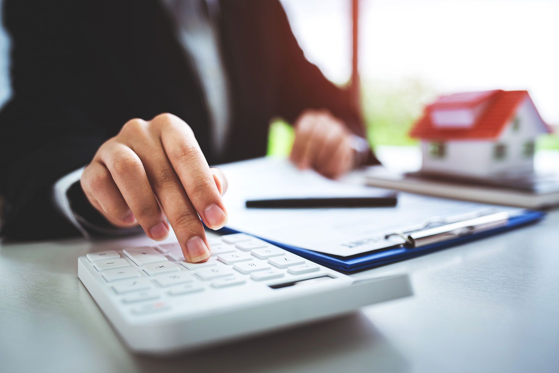 Hand pressing buttons on a white calculator beside paperwork and a pen on a desk
