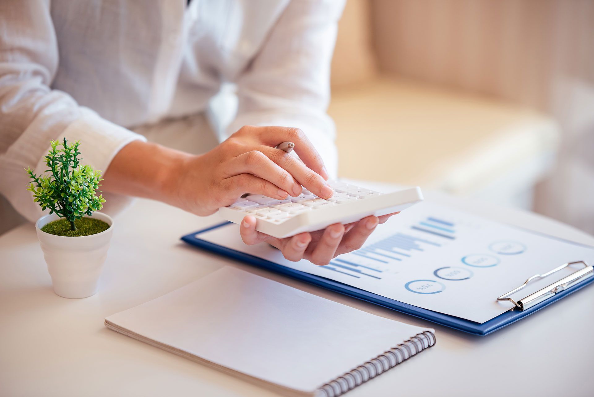 Person entering data on a calculator beside a clipboard, notebook, and small potted plant on a desk.