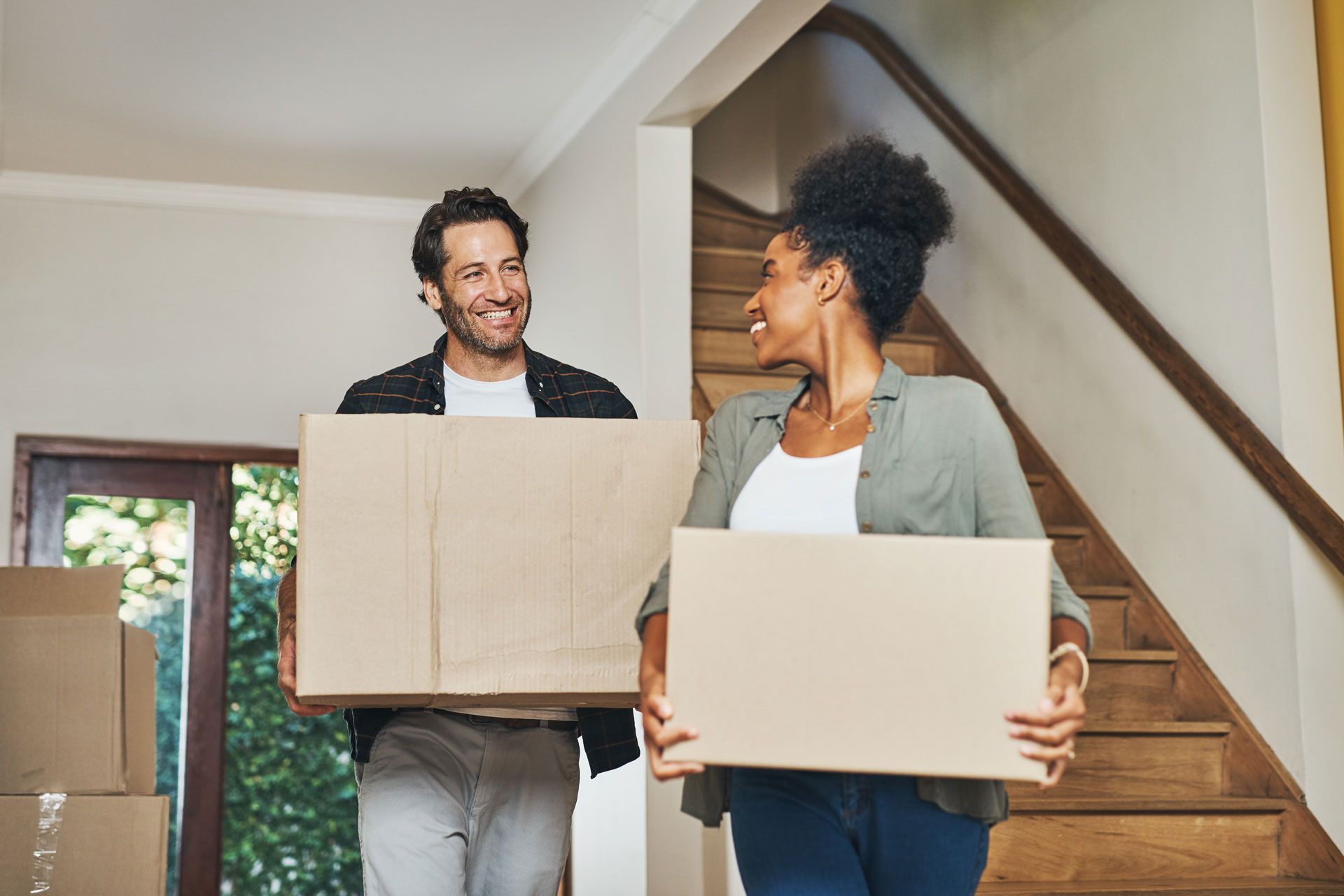 Two people carrying cardboard boxes in a home, smiling on a staircase.