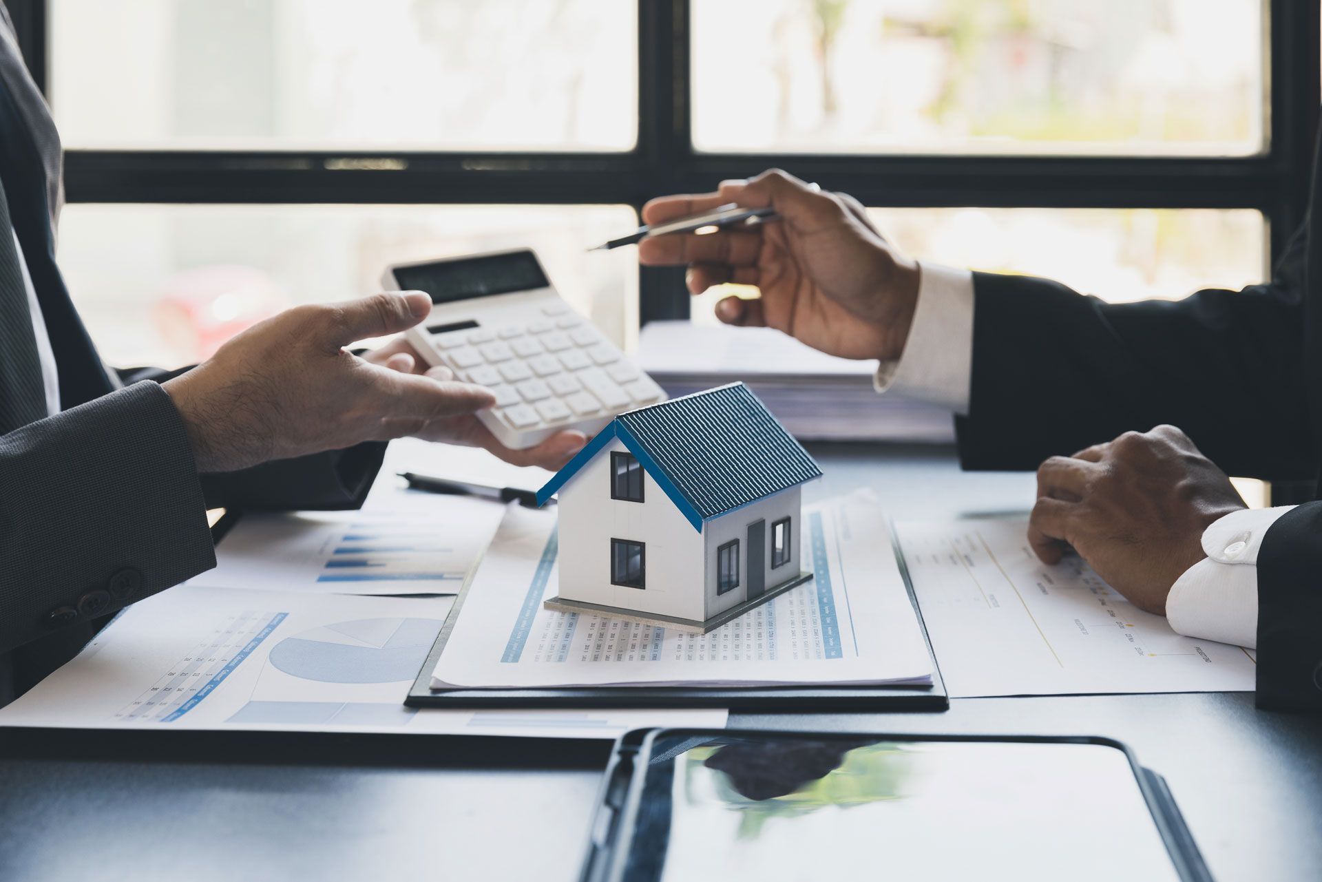 Hands exchanging house keys over a model home and documents on a desk during a real estate meeting