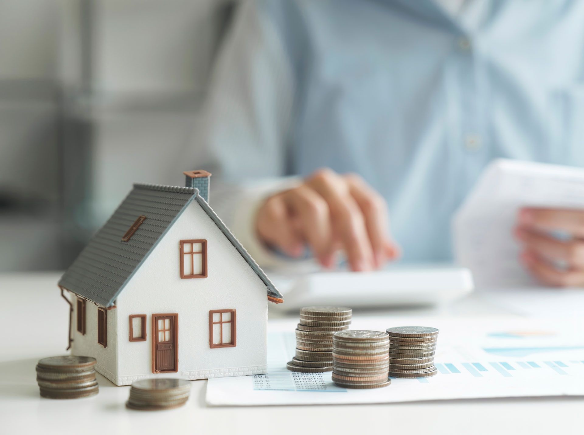 Miniature house with stacked coins and a blurred person in the background, suggesting home savings or mortgage planning