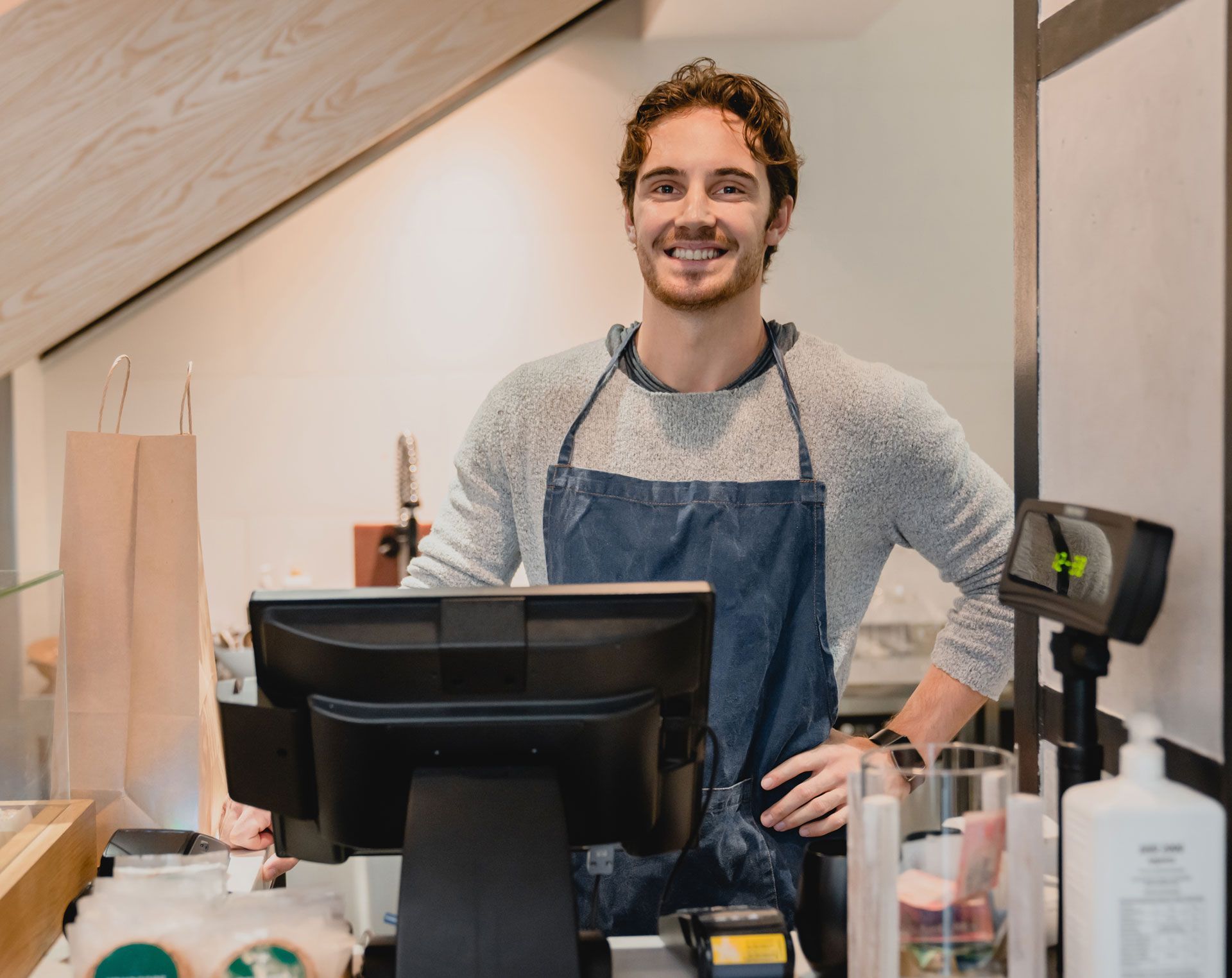 Smiling shop clerk at a checkout counter in an apron, with shelves and retail items behind him