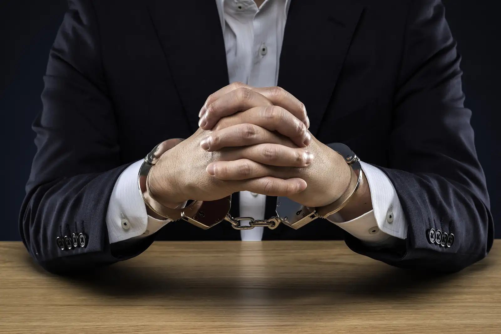 A man in a suit is sitting at a table with his hands in handcuffs.