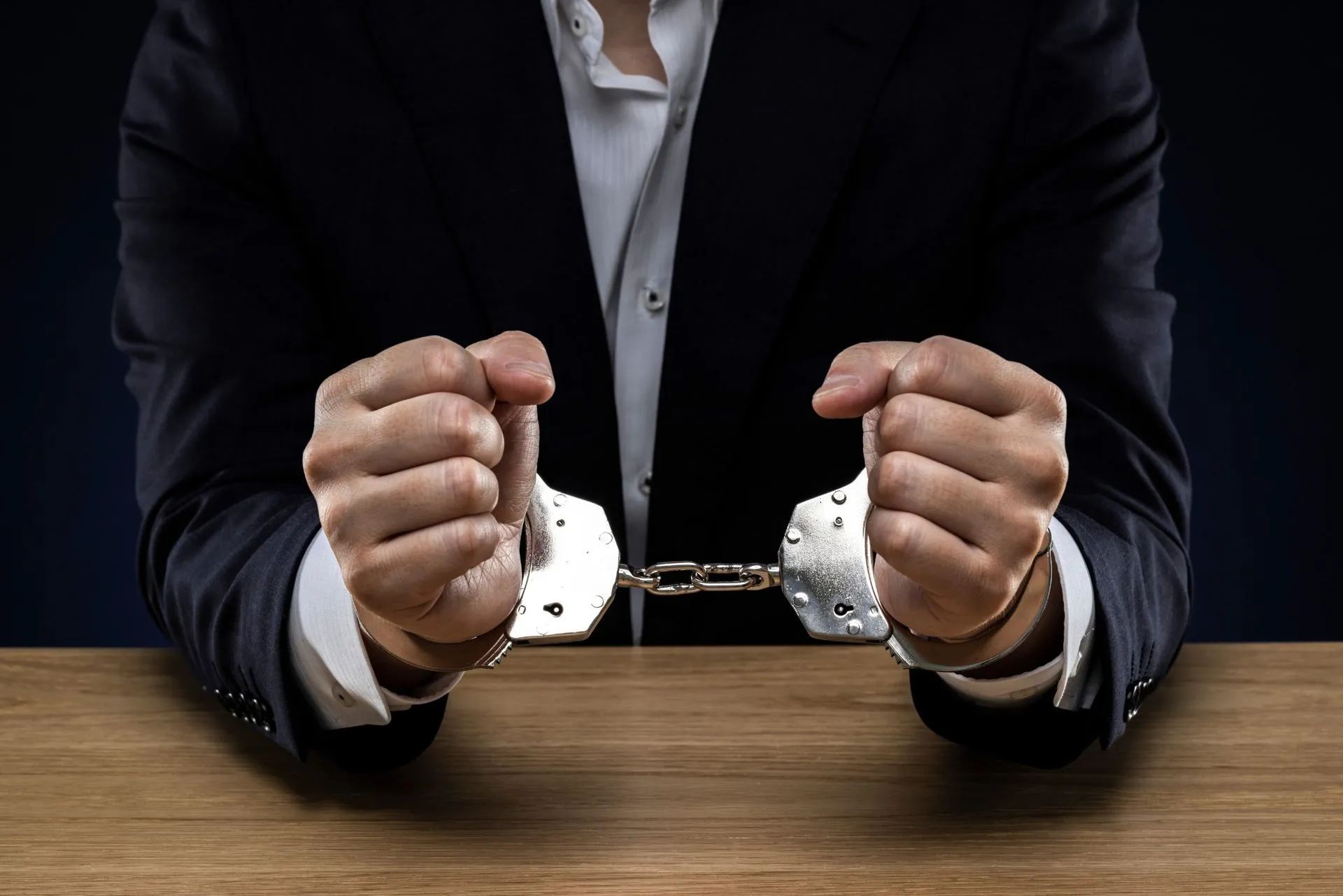 A man in a suit is sitting at a table with his hands in handcuffs.