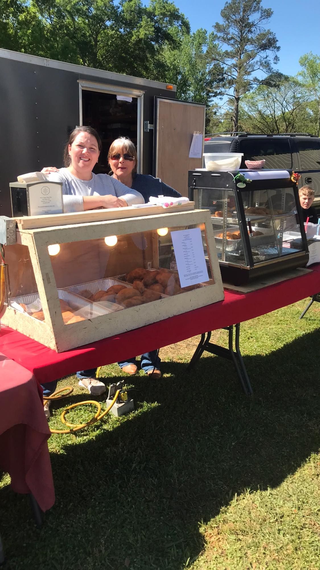 Two people at a food stall selling baked goods. Display cases on a red table, white trailer in the background.