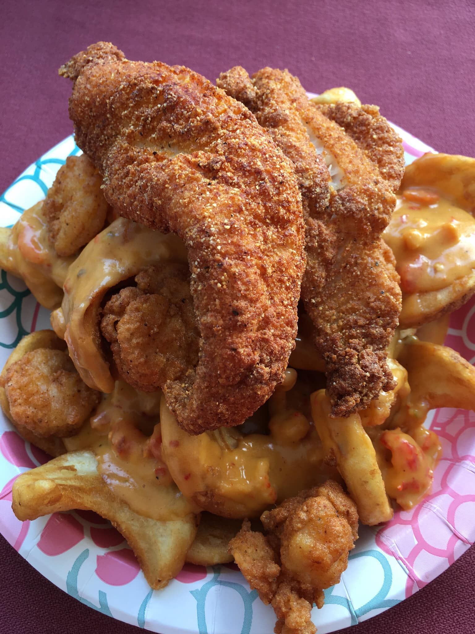 Fried fish and shrimp atop fries, drizzled with sauce on a paper plate.