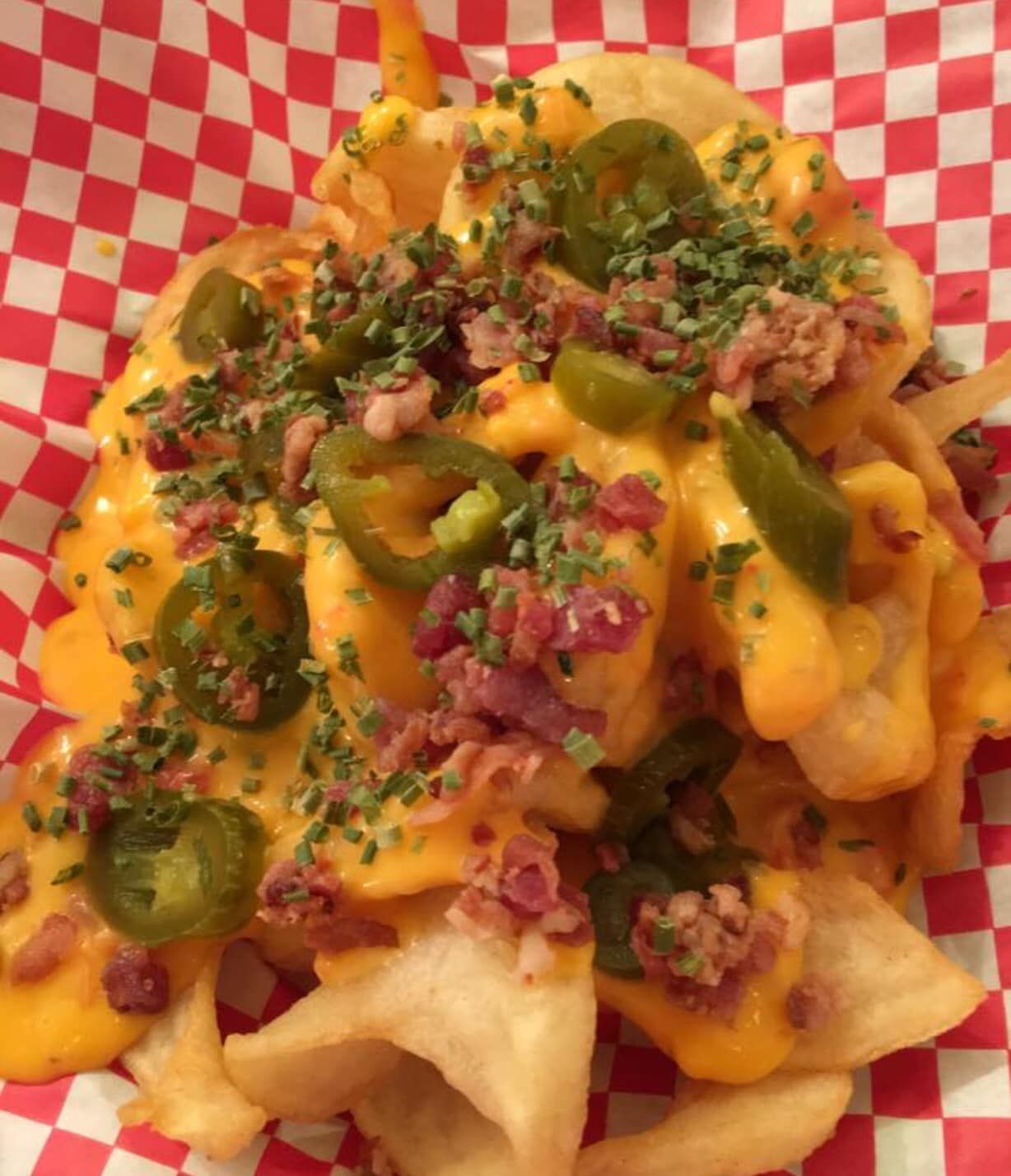 Fries topped with melted cheddar cheese, bacon, jalapeños, and chives in a red and white checkered paper.
