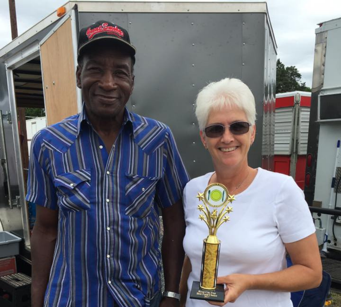 Man and woman standing next to a trailer, woman holding a trophy. Both smiling.
