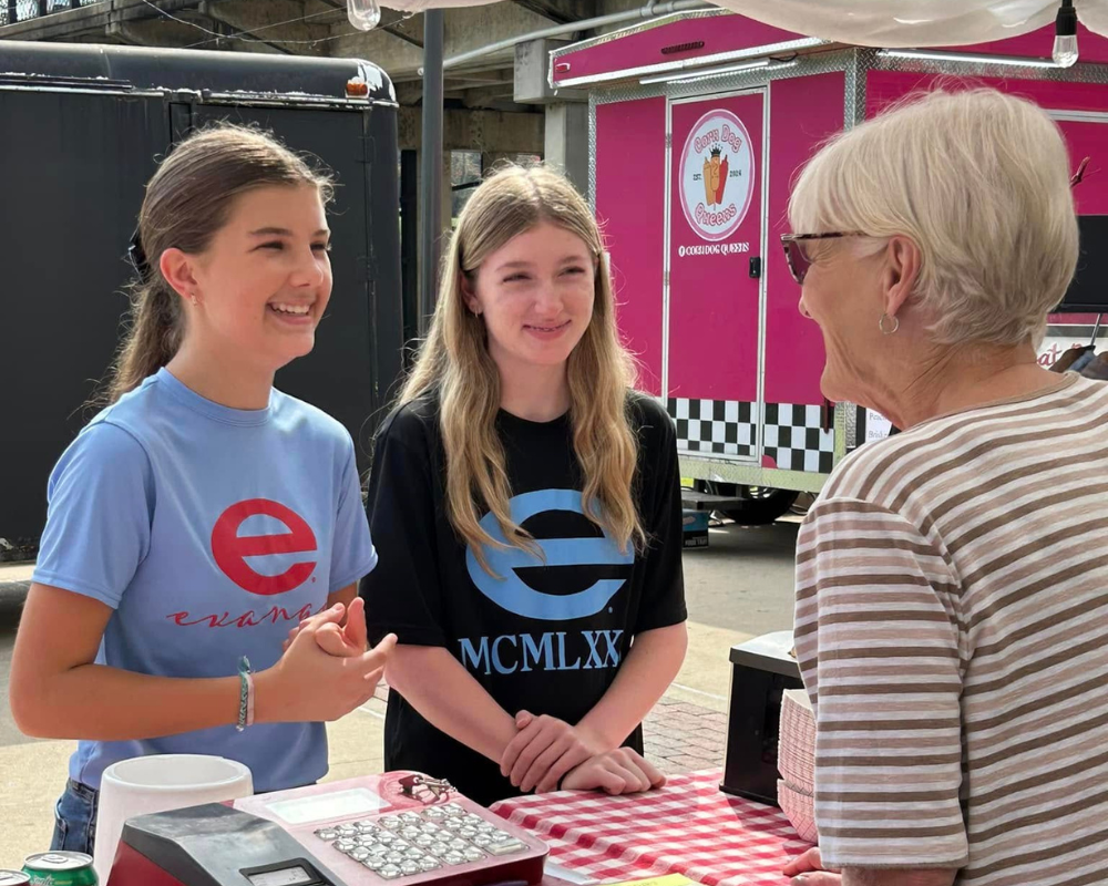 Two young people selling ice cream from a food truck, talking with a customer at a red table.