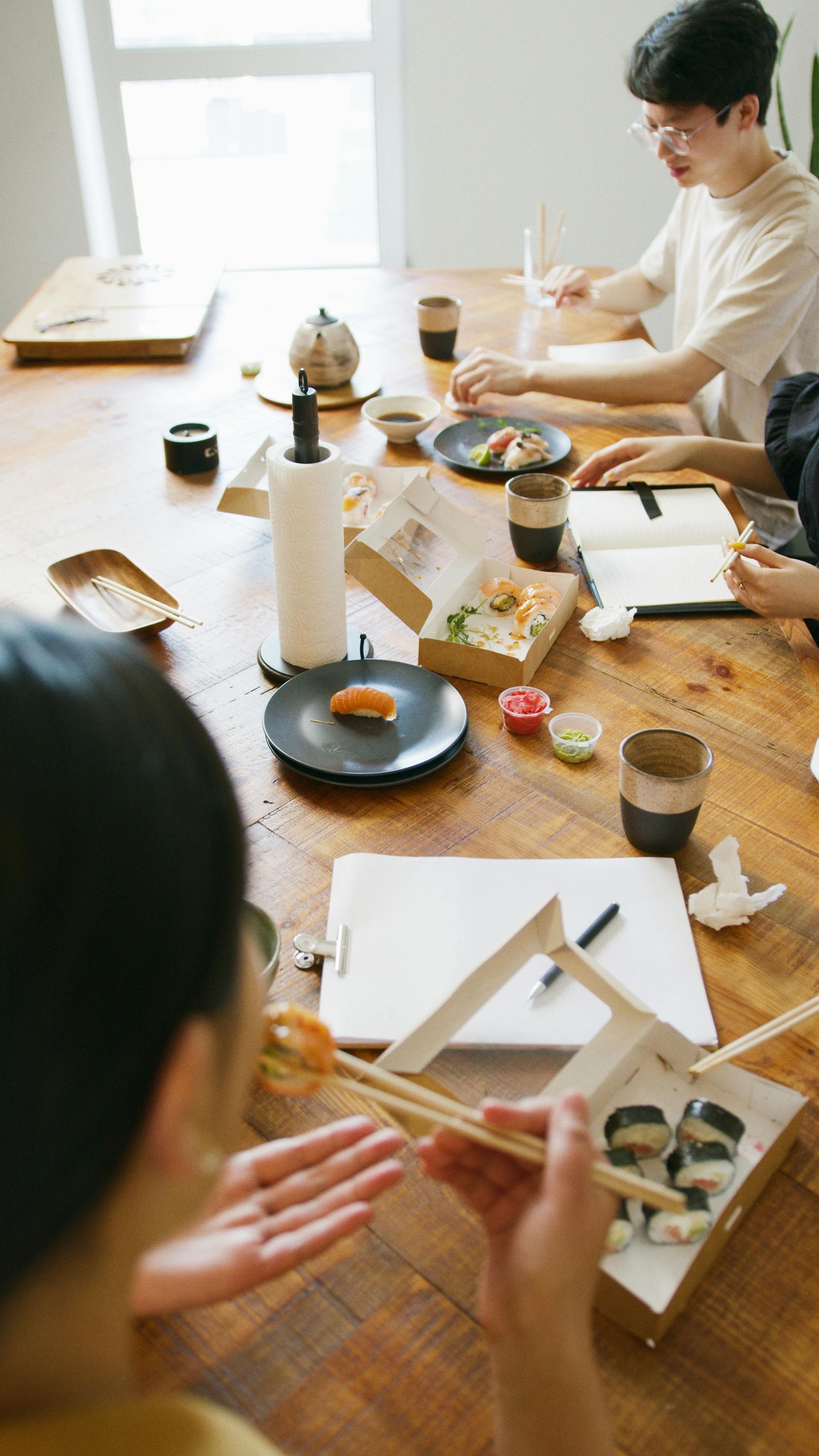 People eating sushi at a wooden table, using chopsticks. Some have open food boxes and drinks.