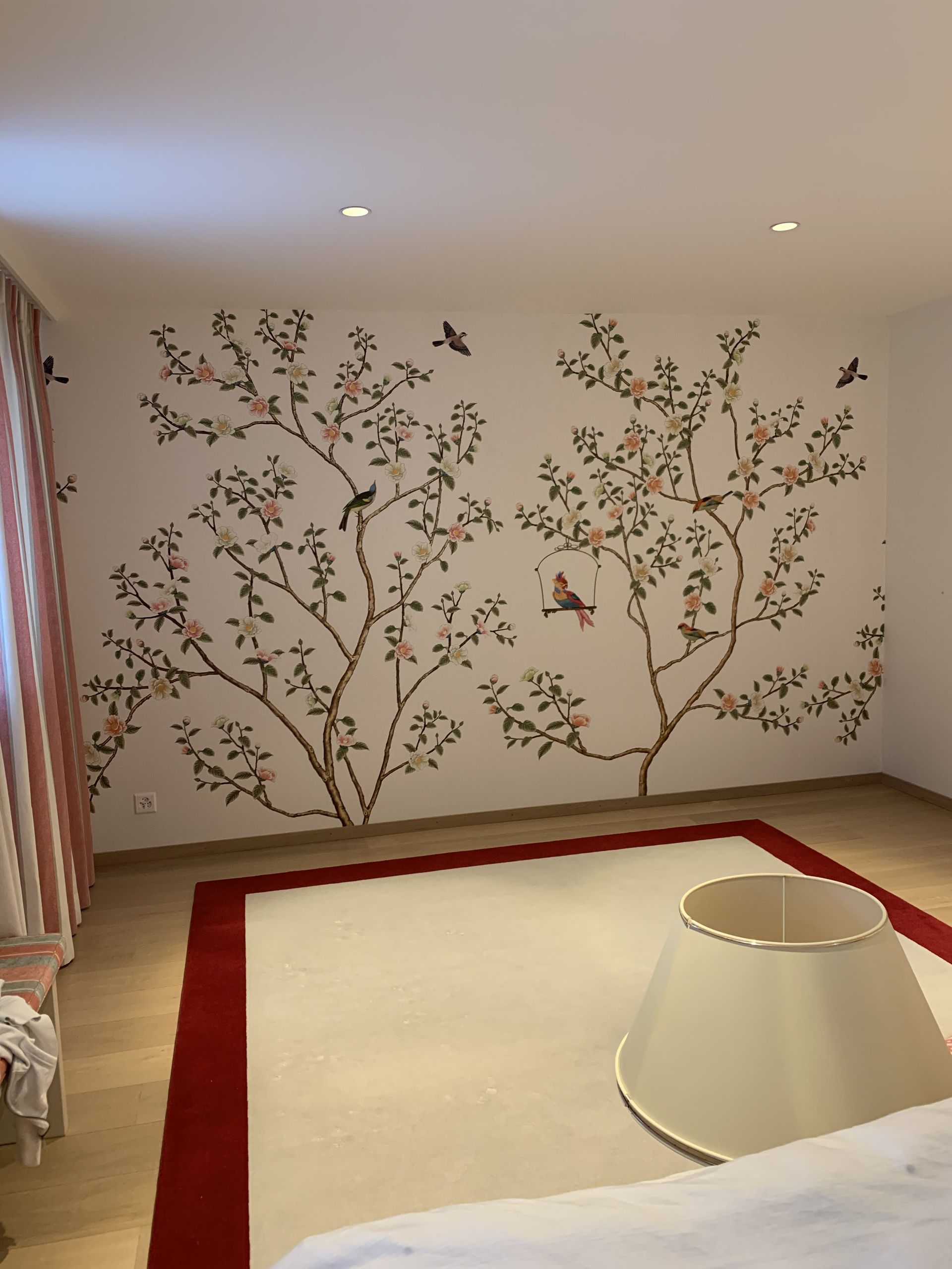Une petite pièce avec des stickers muraux à motifs floraux, un tapis crème à bordure rouge et un abat-jour blanc posé au sol.
