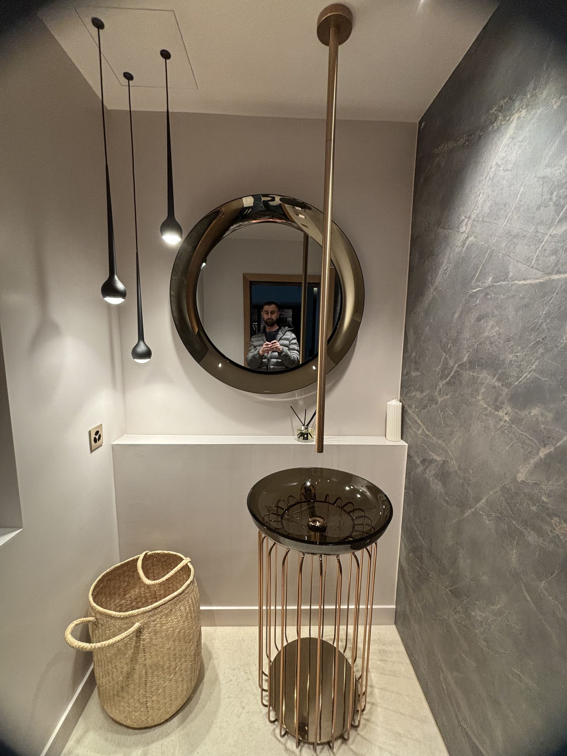 Salle de bains moderne avec suspensions lumineuses, miroir rond, robinetterie en laiton, paniers tressés et lavabo sur plan de travail noir