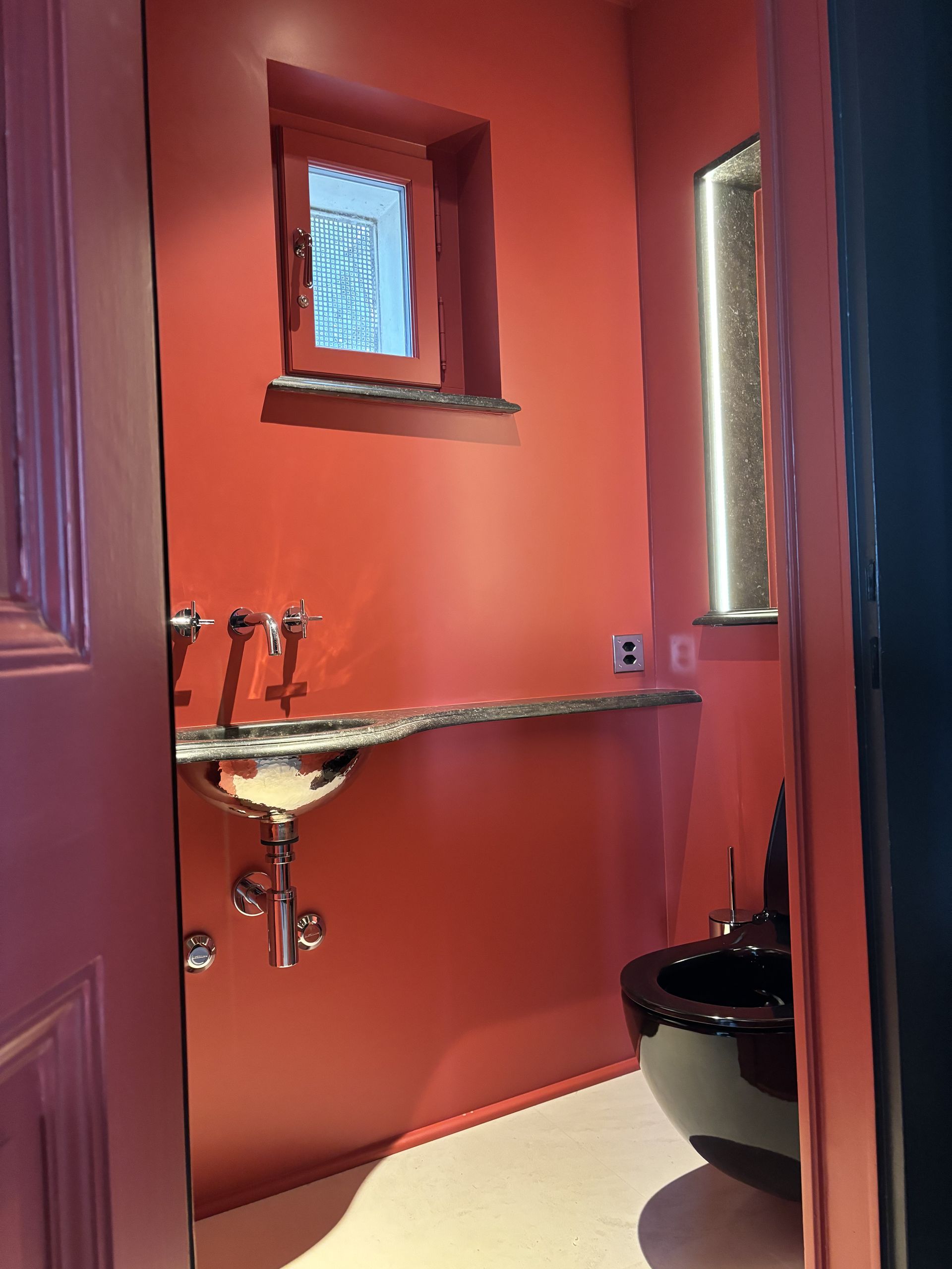 Petite salle de bains aux murs corail, lavabo sur pied, miroir et toilettes noires partiellement visibles.