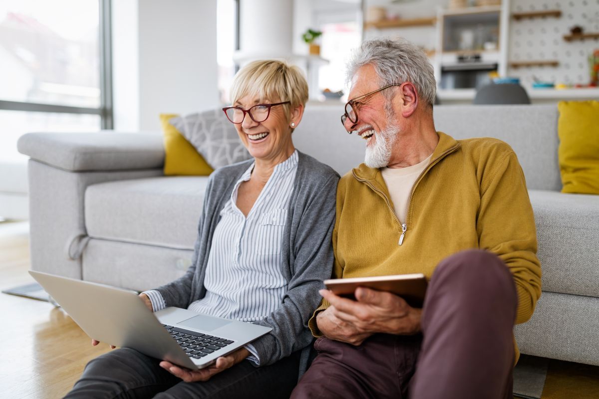 An elderly couple is sitting on the floor using a laptop and a tablet.