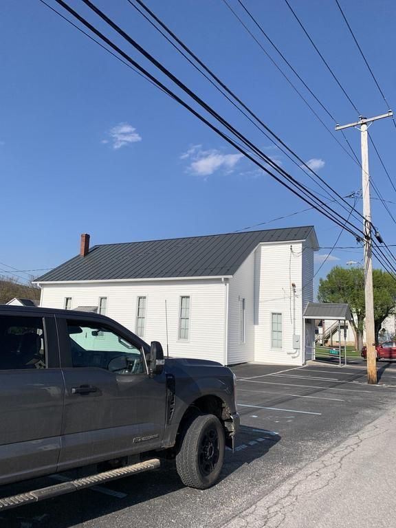 A gray truck parked in front of a white building with a dark gray roof. Overhead, power lines are strung against a blue sky.