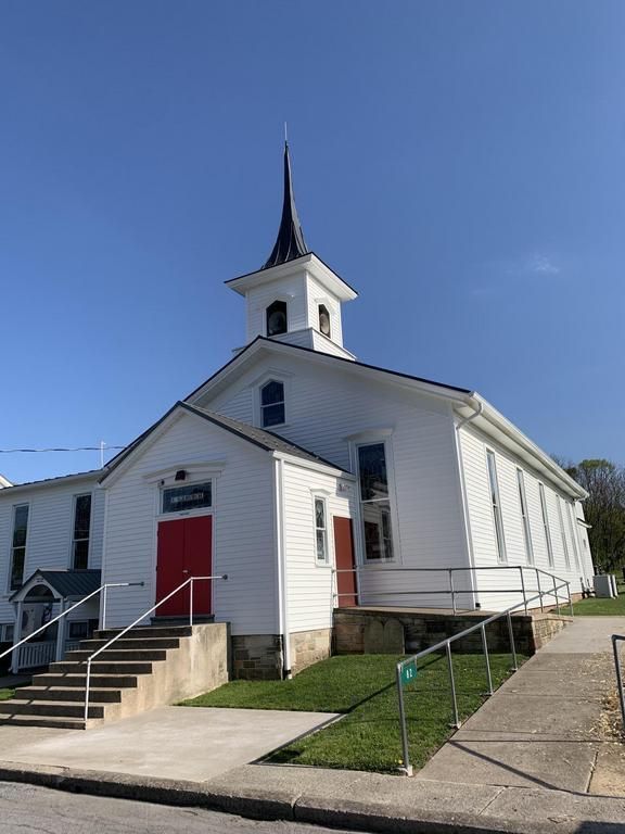 White church with a red door, steeple, and concrete steps leading up to it. The sky is blue.