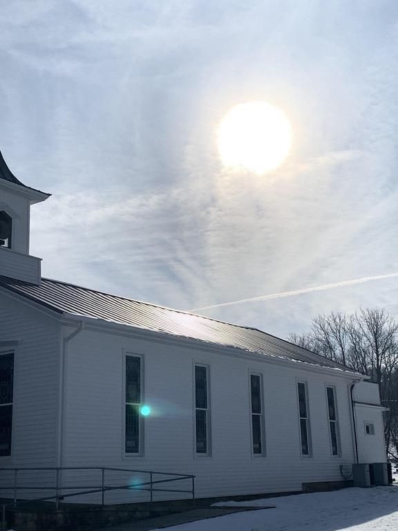 White church with a bell tower, side profile, under a bright sun and cloudy sky. Snow on the ground.