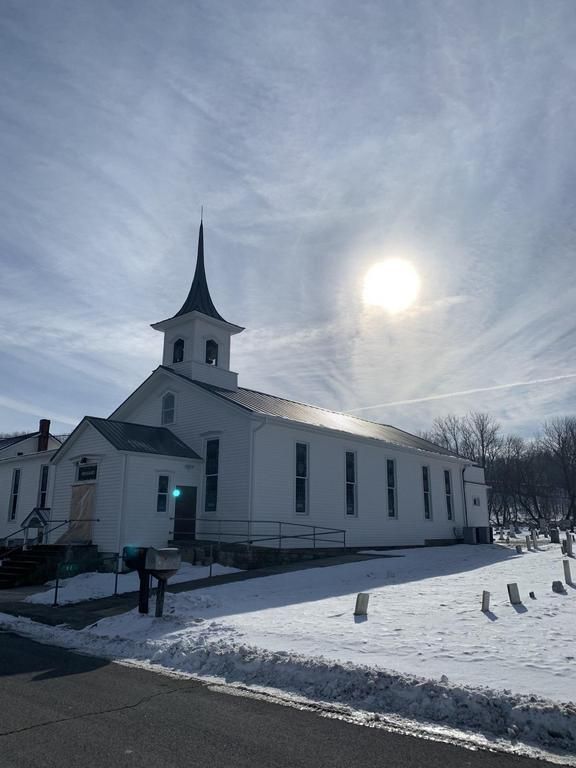 White church with a steeple against a sunny winter sky; snow covers the ground and a cemetery is visible to the side.