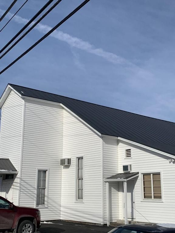 White building with dark gray roof against a blue sky with a contrail. Power lines are in the foreground.