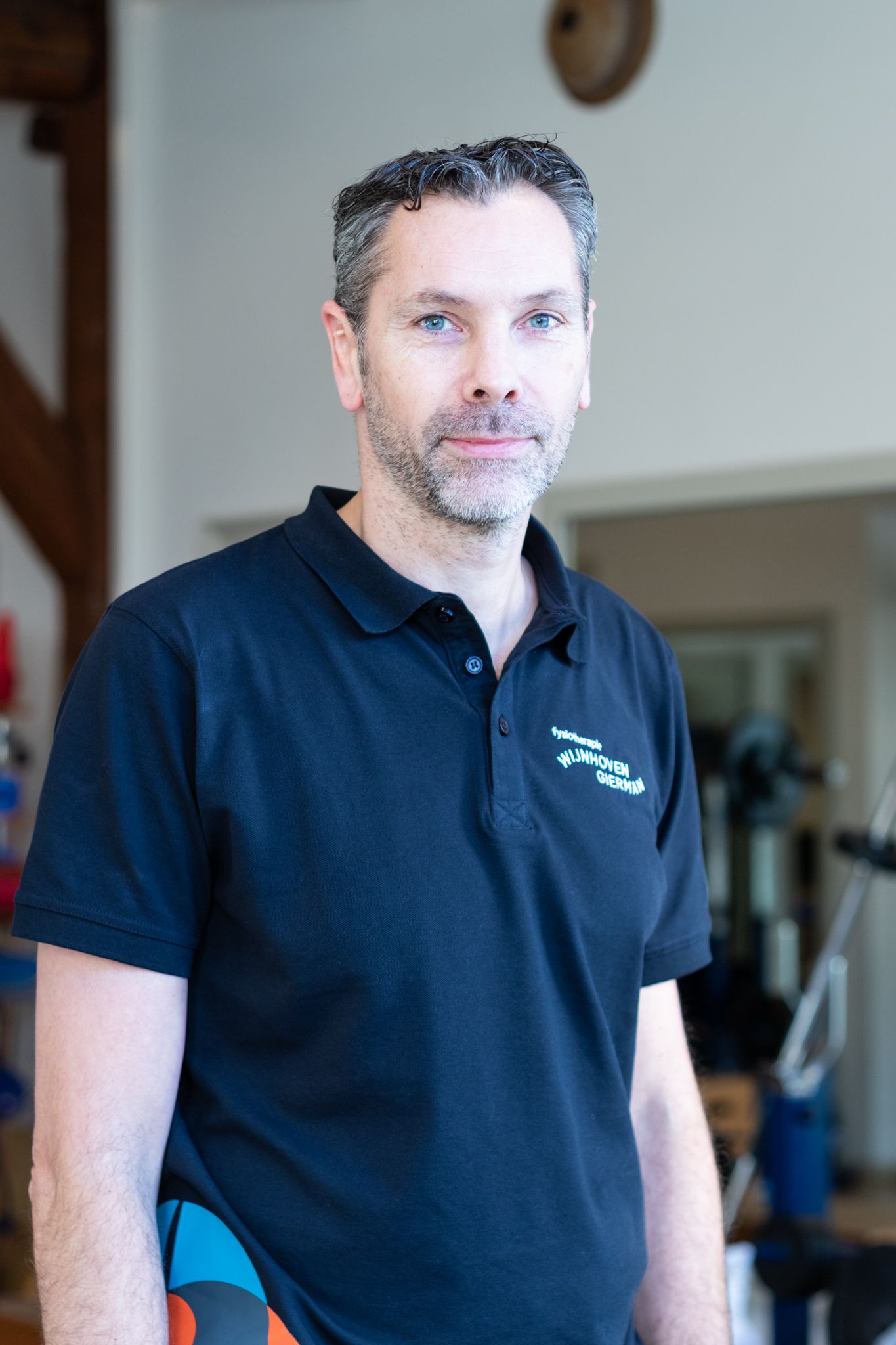 Een man met een baard, gekleed in een zwart poloshirt, staat in een kamer.