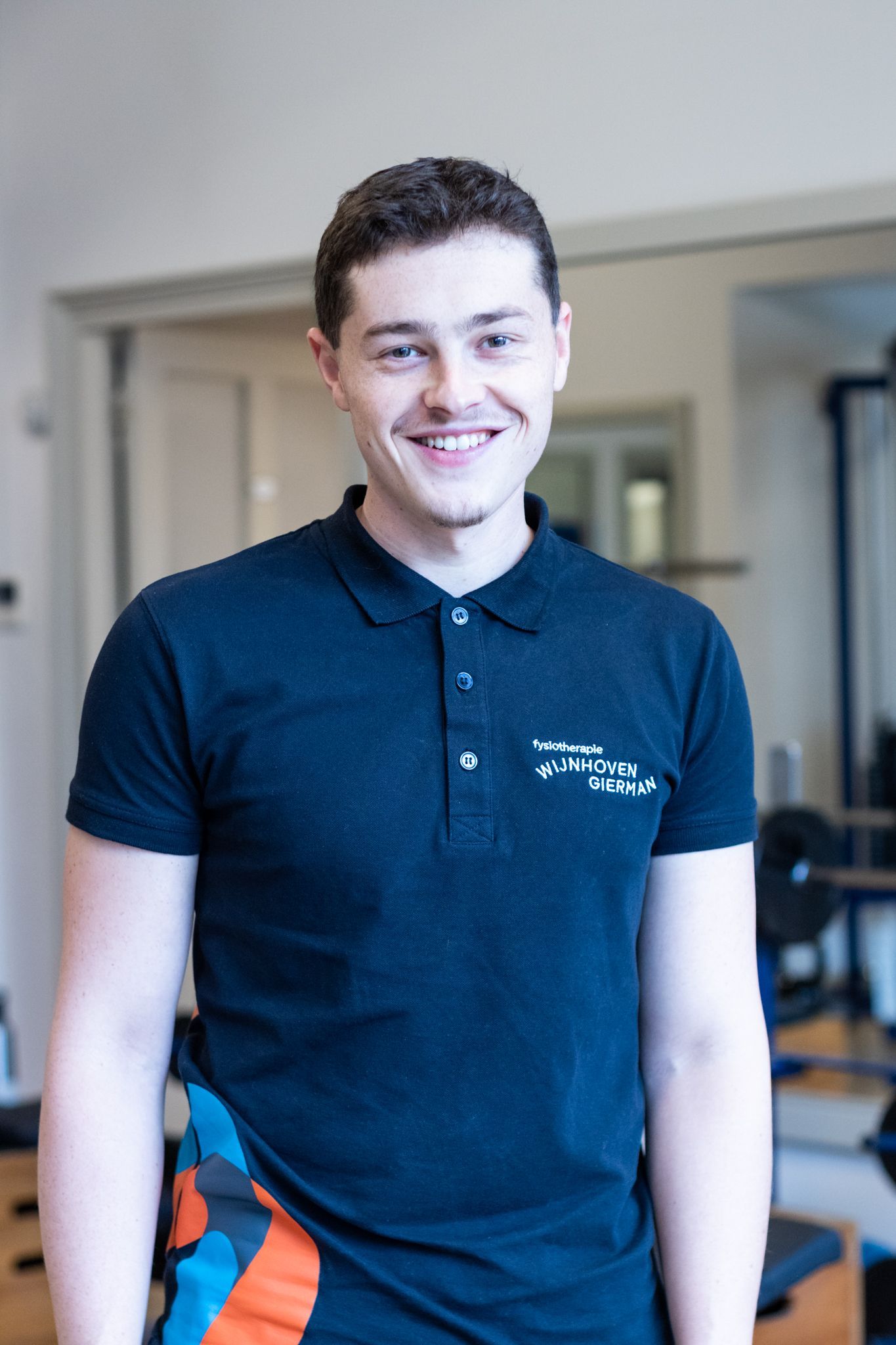 Een jonge man in een zwart poloshirt lacht in een sportschool.