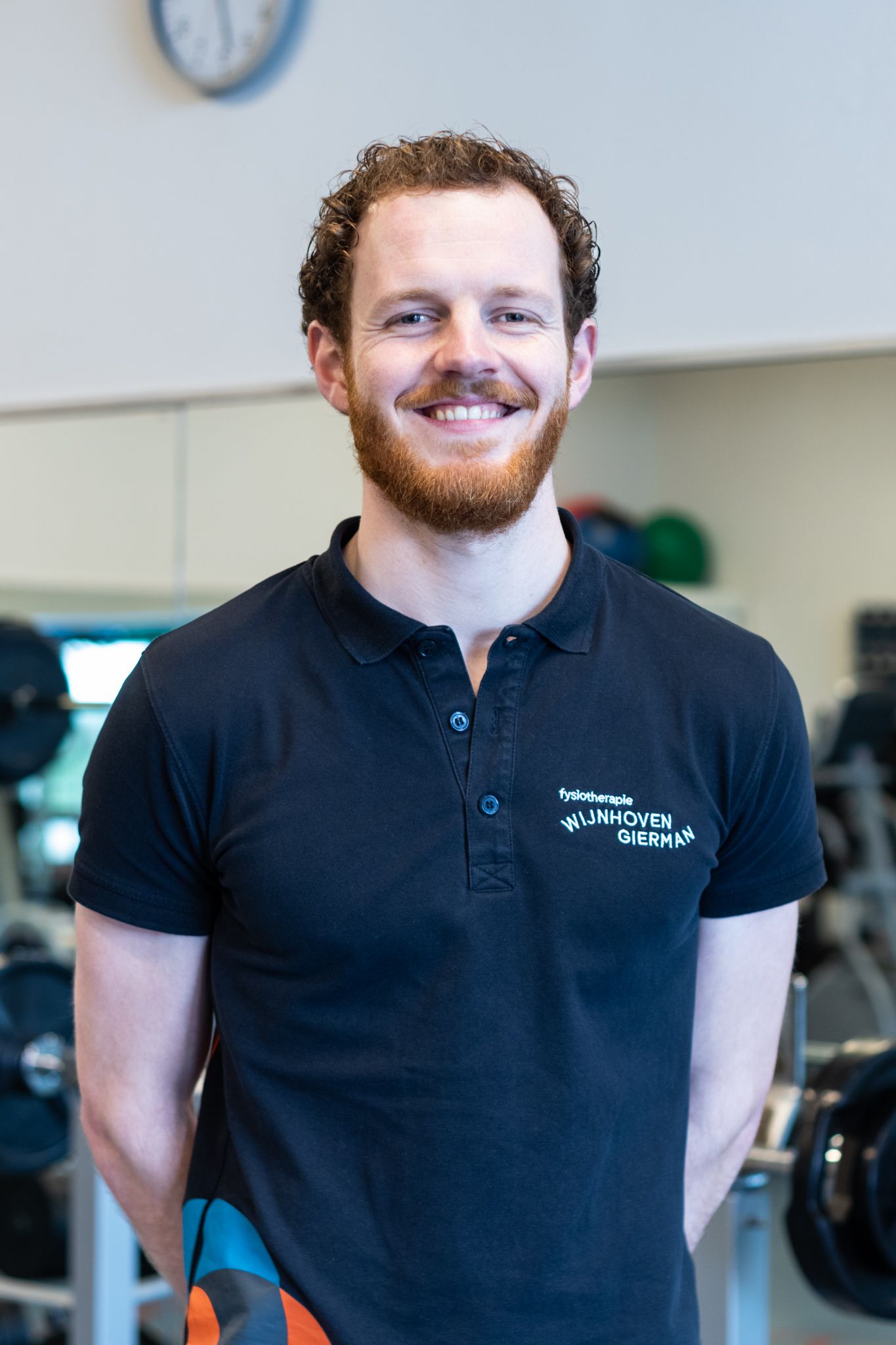 Een man met een baard, gekleed in een zwart poloshirt, glimlacht in een sportschool.