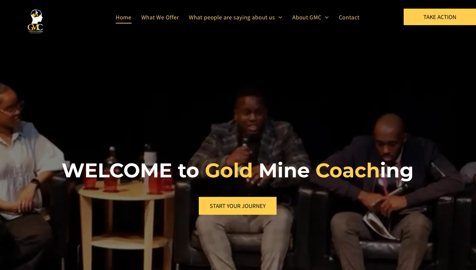 Three men on stage: one speaking with a microphone, two others seated. Gold Mine Coaching website.