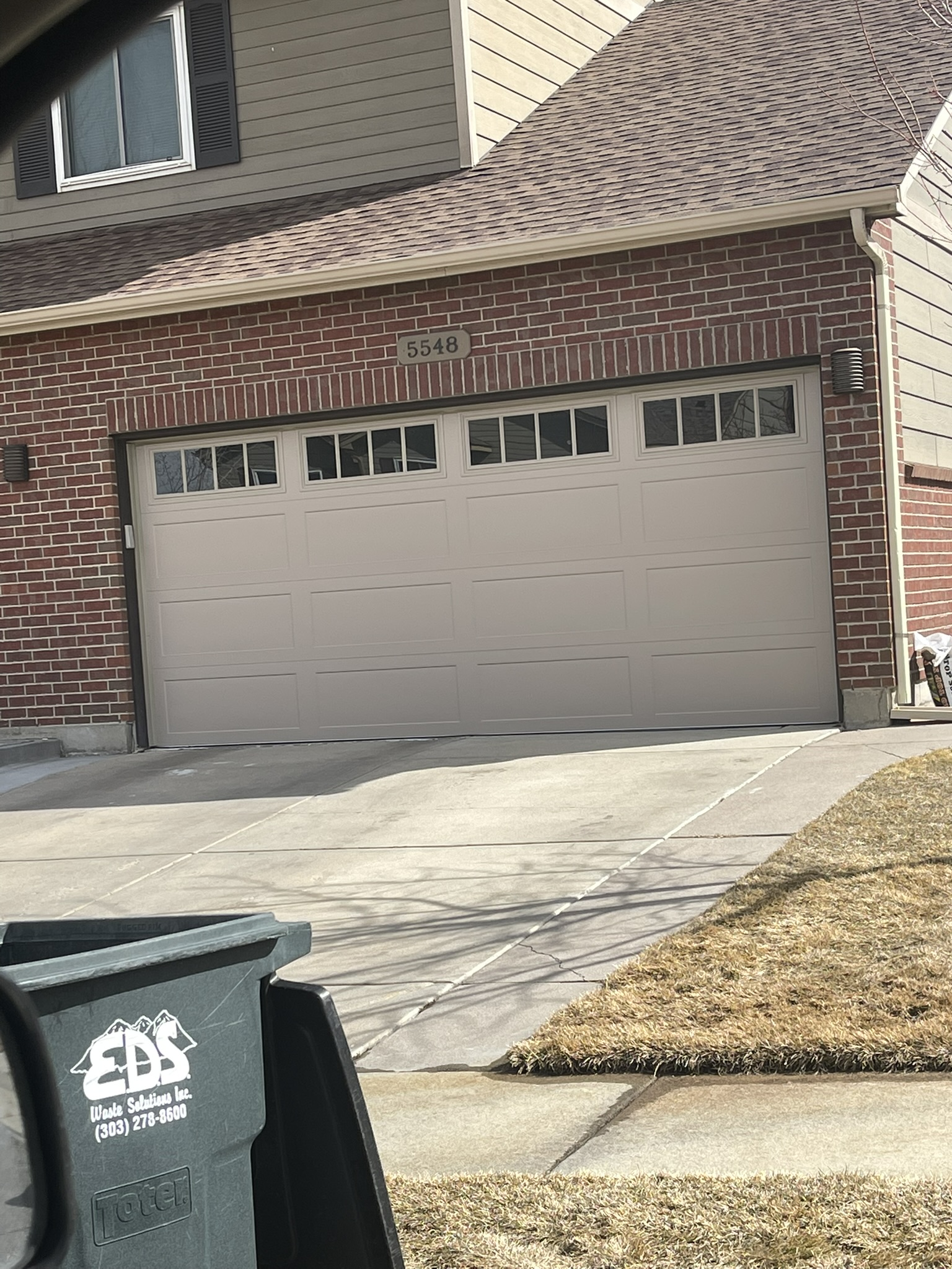 A brick house with a garage door and a trash can in front of it.