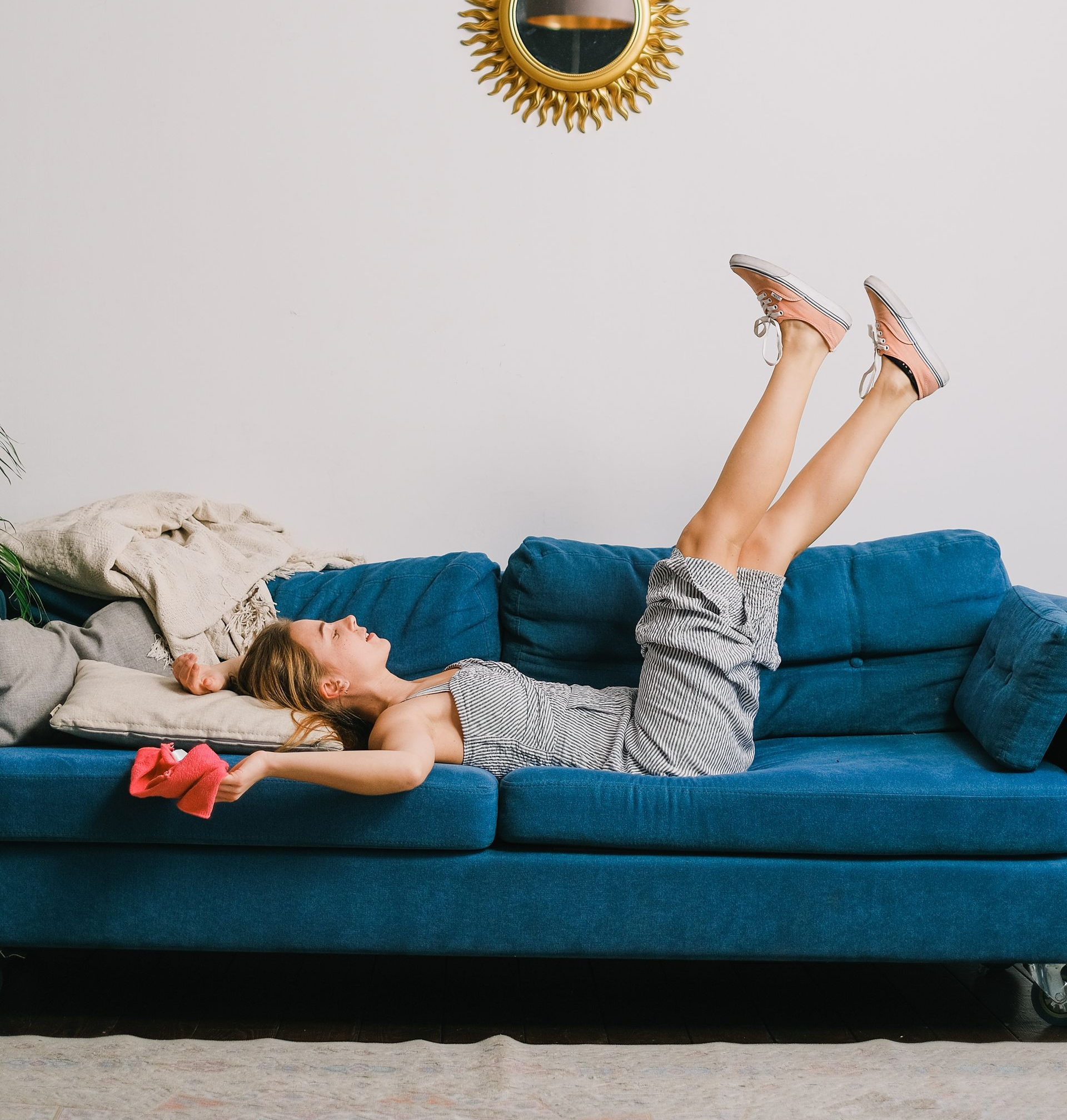 Person lying on a blue couch with legs raised, in a bright living room with a gold sunburst mirror.