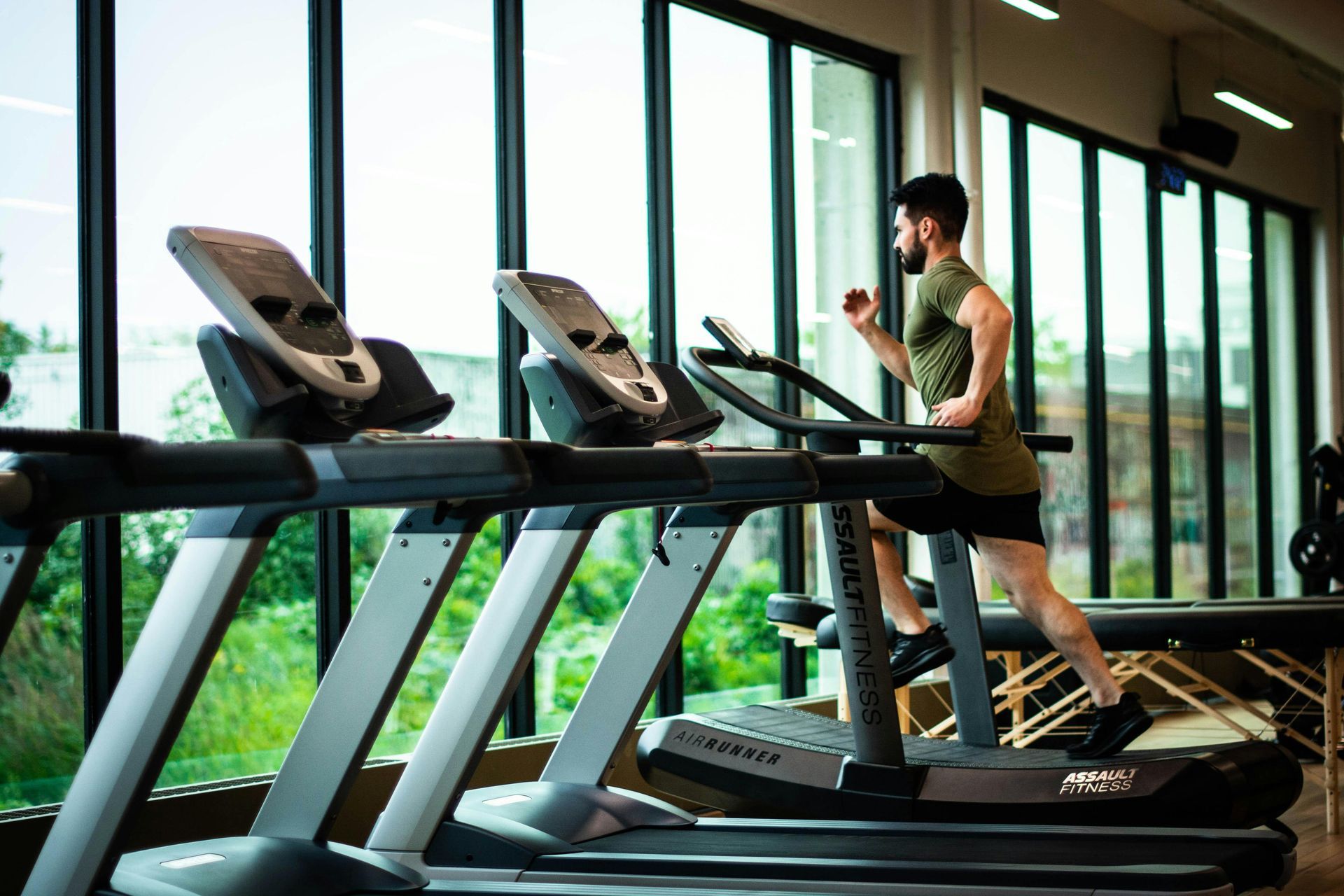 A person wearing an olive green t-shirt and dark shorts runs on a treadmill in a gym with large floor-to-ceiling windows.