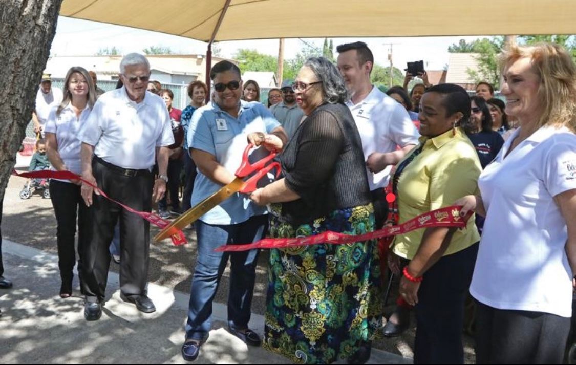 Man hands a document to a woman during a ribbon-cutting ceremony under a canopy; crowd applauds.