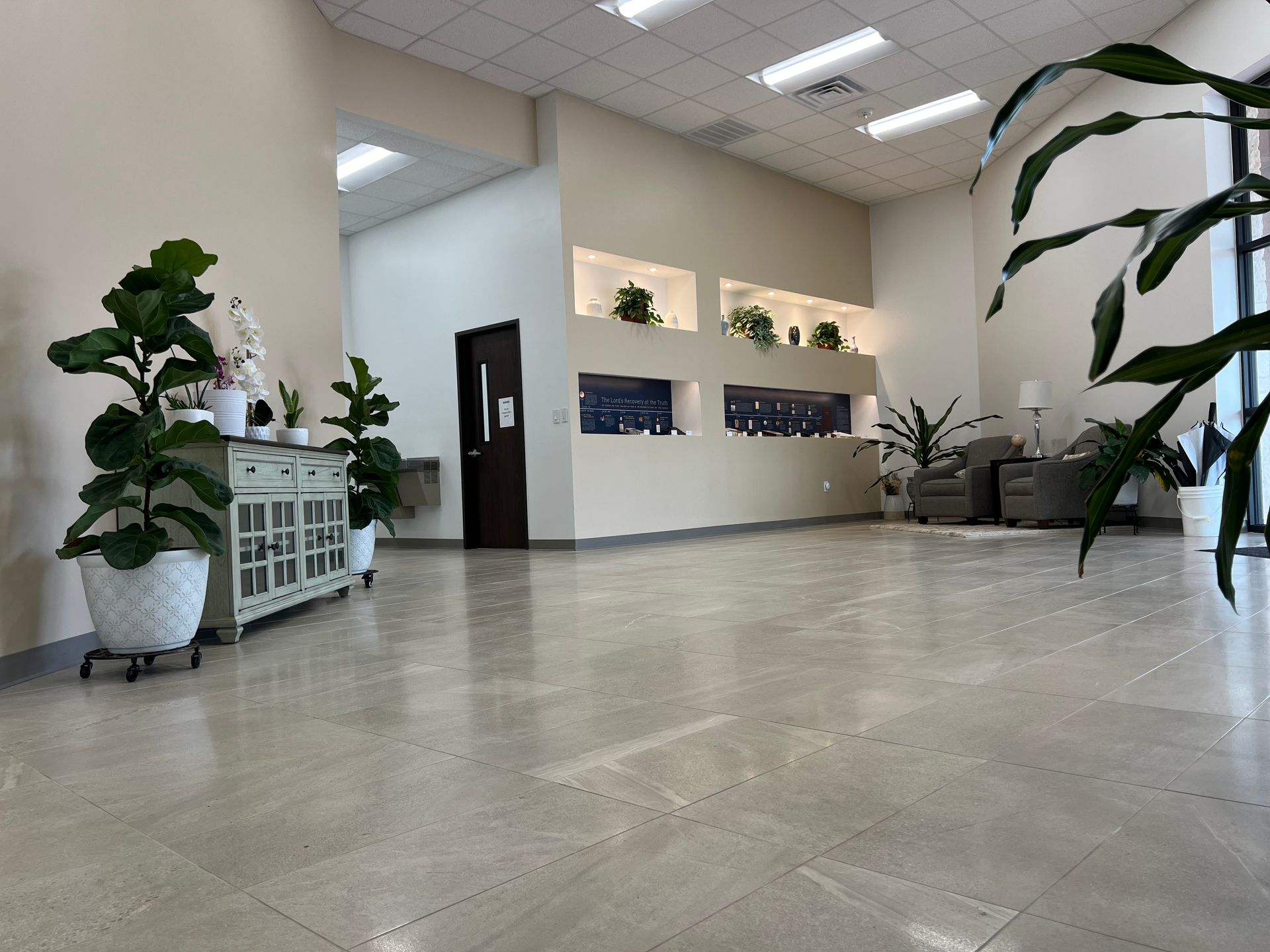 Spacious waiting area with plants, built-in wall displays, beige tile floor, and seating area.