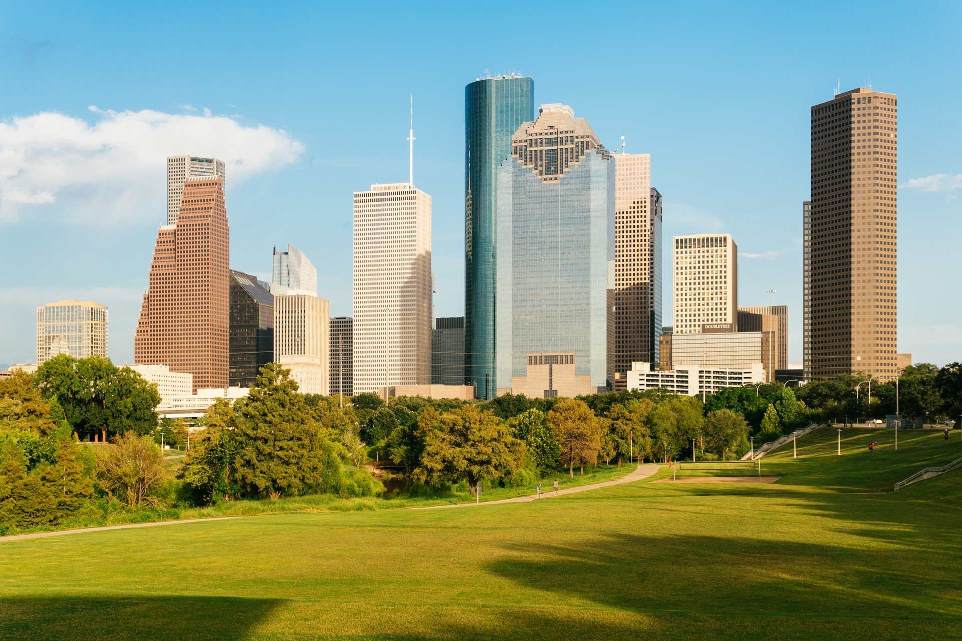 Houston skyline with skyscrapers, green trees, and a grassy park under a blue sky.