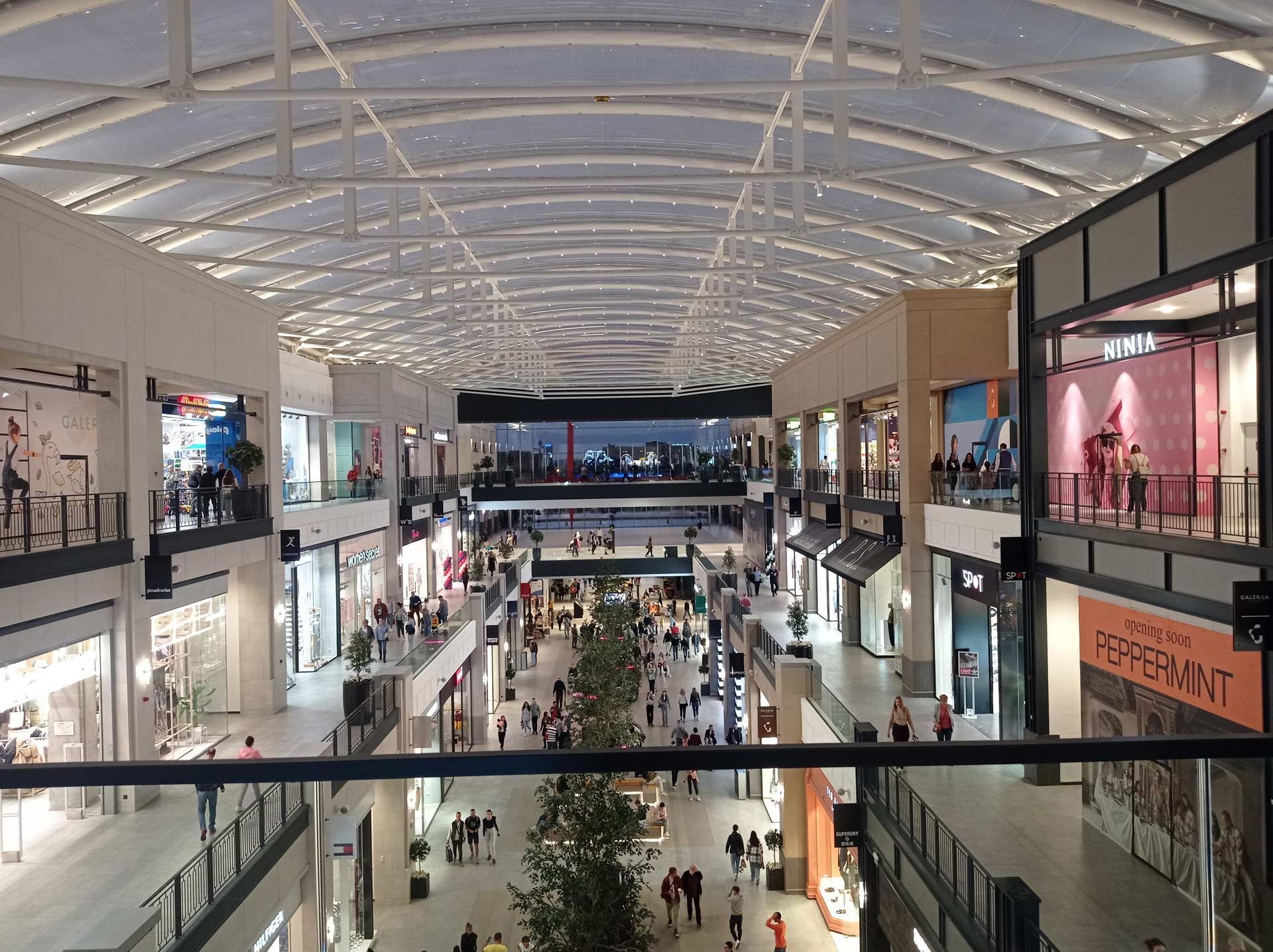 Interior view of a multi-story shopping mall with shoppers, stores, and a curved glass ceiling.