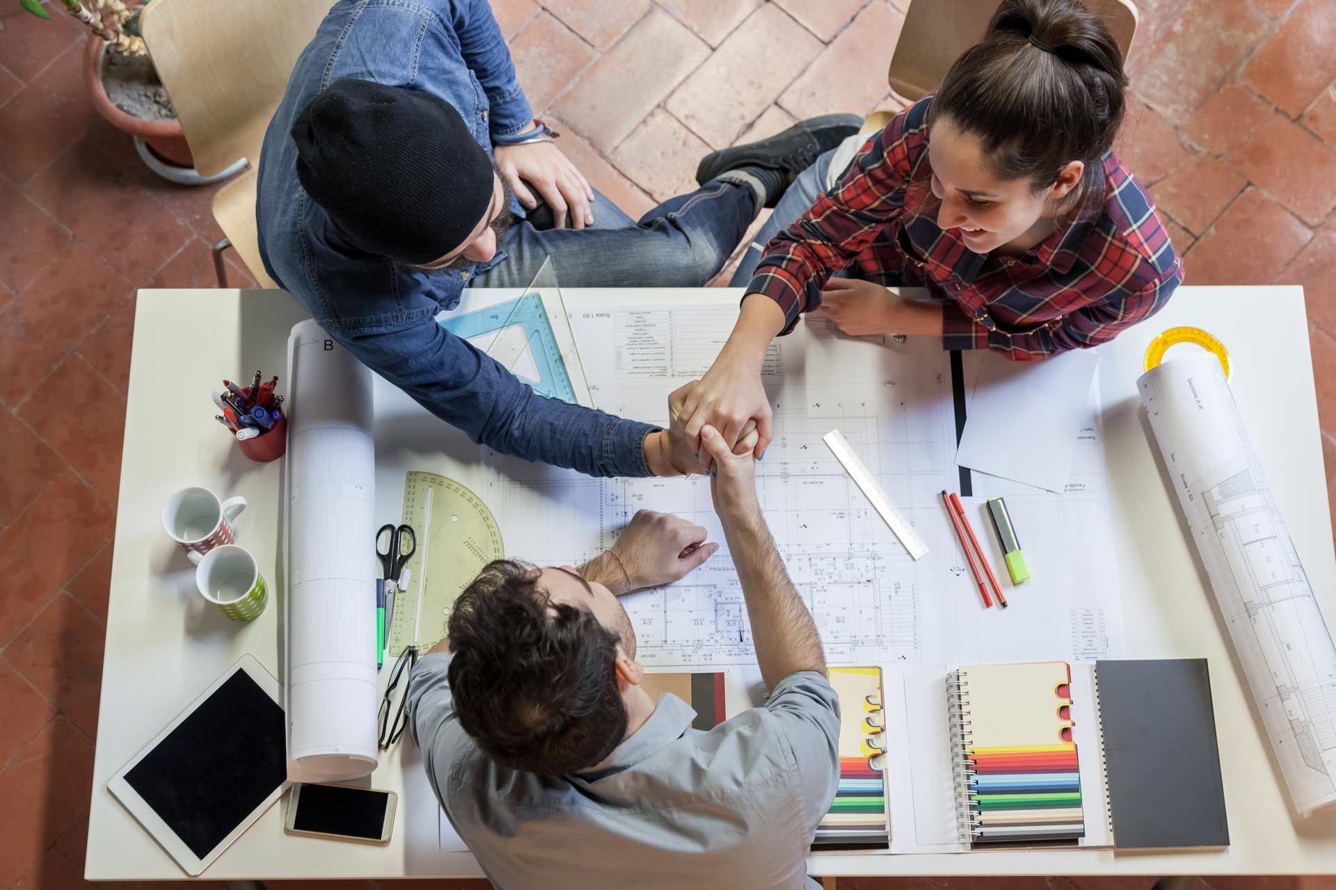 Three people fist-bumping over architectural plans on a table.