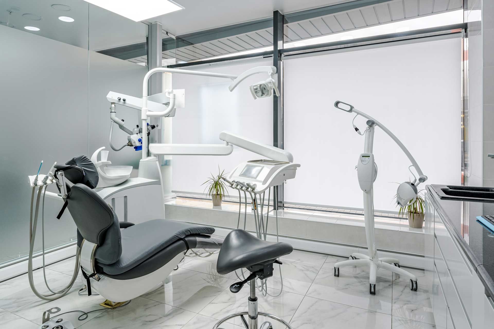 Dental office interior with chair, equipment, and large window with shades.