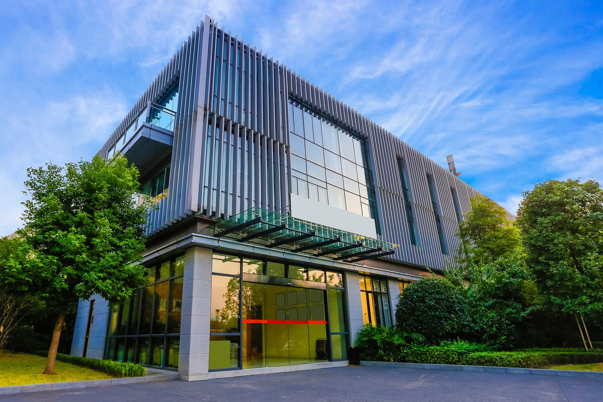 Modern building with glass facade, vertical gray stripes, and surrounding green trees under a blue sky.