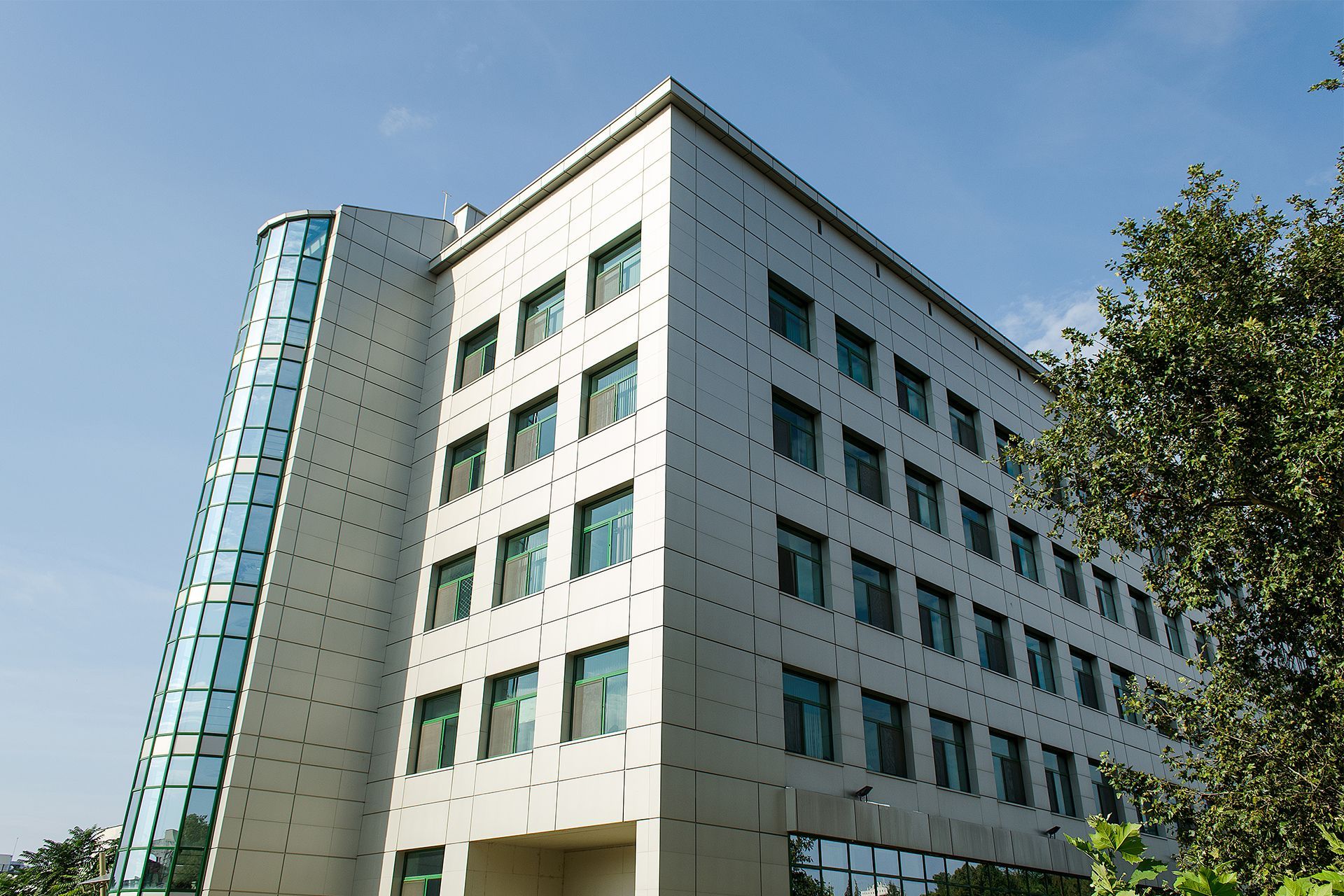 Modern building with silver facade, glass tower, and green-trimmed windows under blue sky.