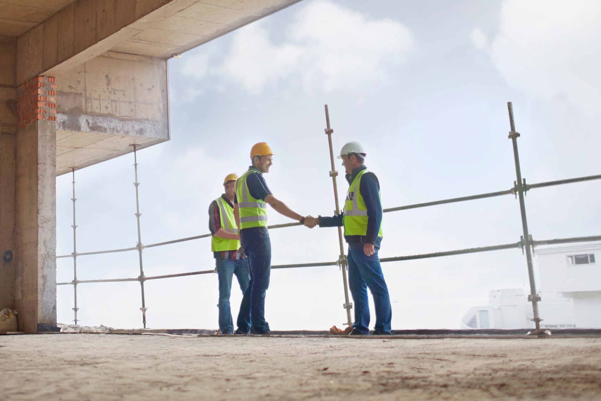 Construction workers shaking hands on a building site; two wear yellow vests and hard hats.