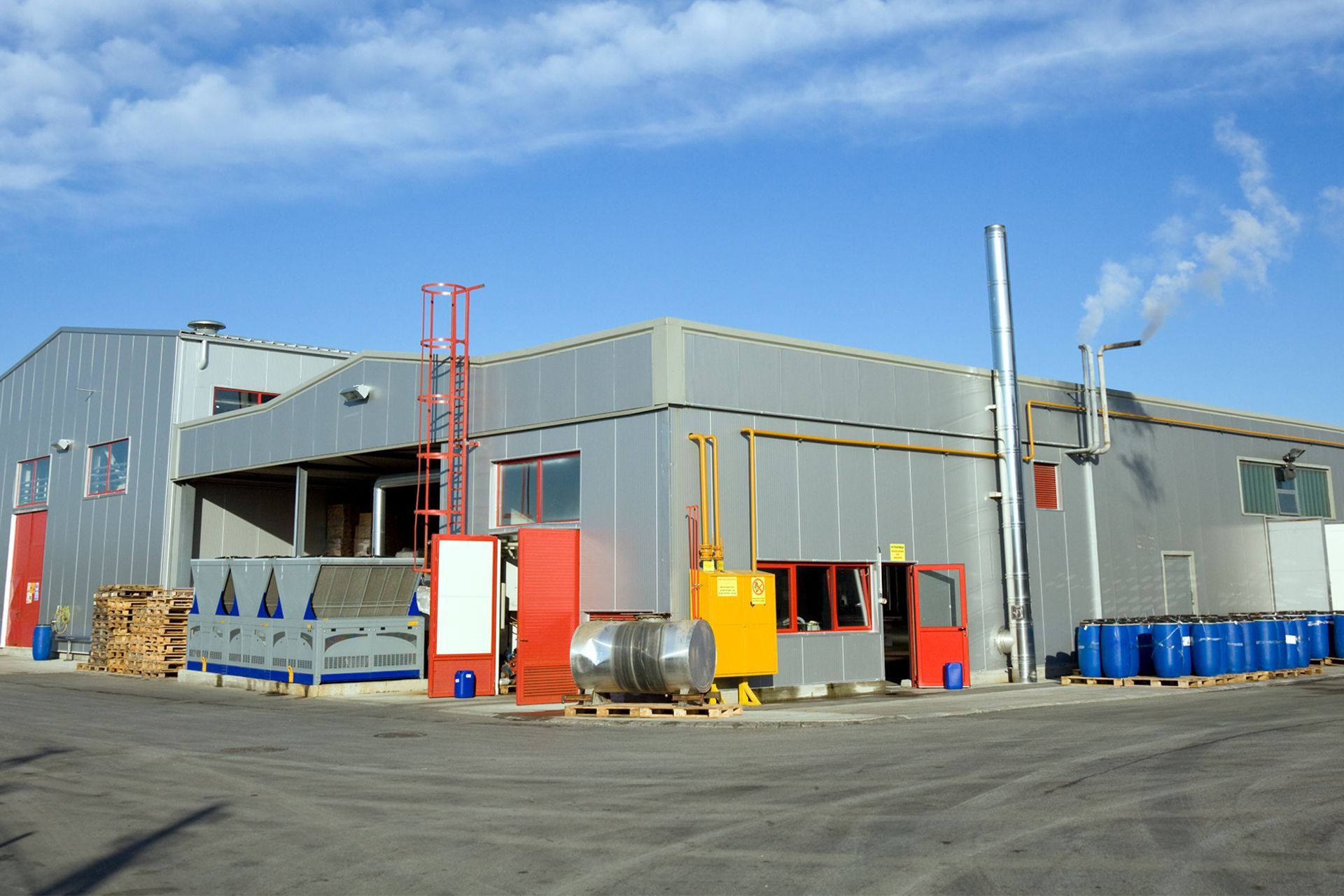 Industrial building, exterior view, metallic siding, red doors, blue barrels, sunny day.